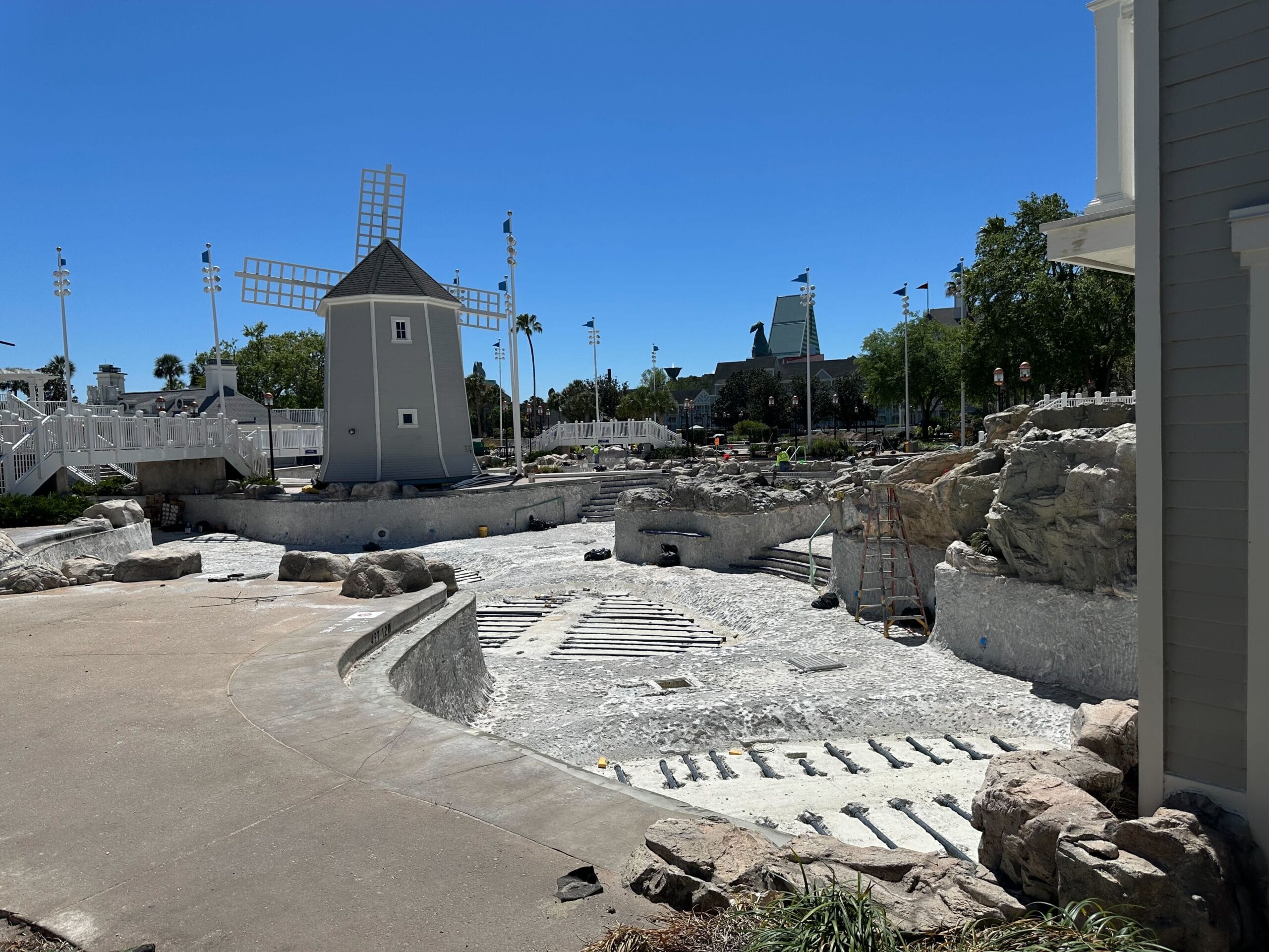 The construction site at Disney's Yacht Club features an empty Stormalong Bay Pool, a ladder, and a windmill structure under a clear blue sky. Rocks and unfinished surfaces are visible in the fenced-off area.