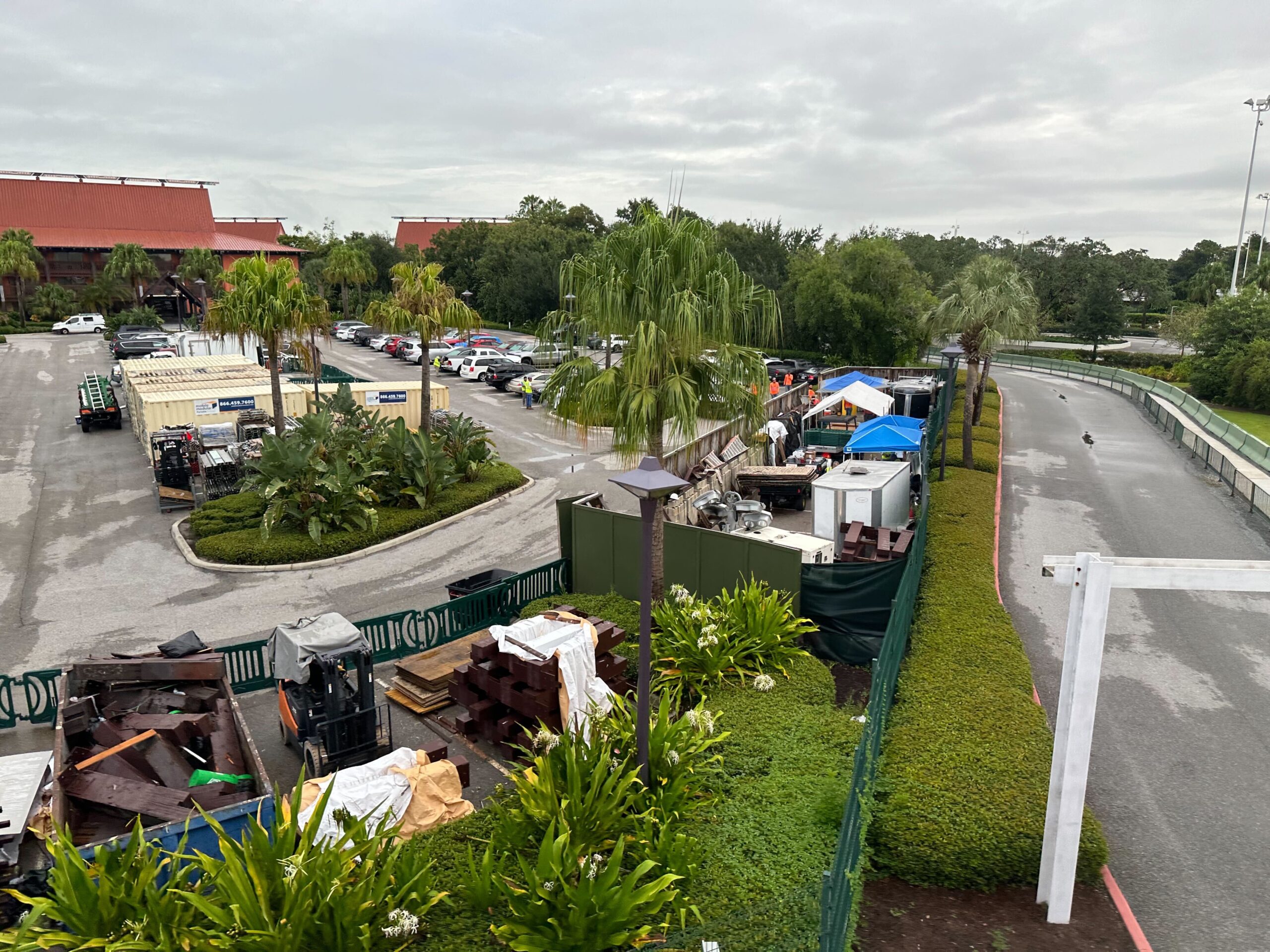 A construction site with equipment and materials is situated near a parking lot and palm trees. Several tents and vehicles are present, and a partially cloudy sky is visible.