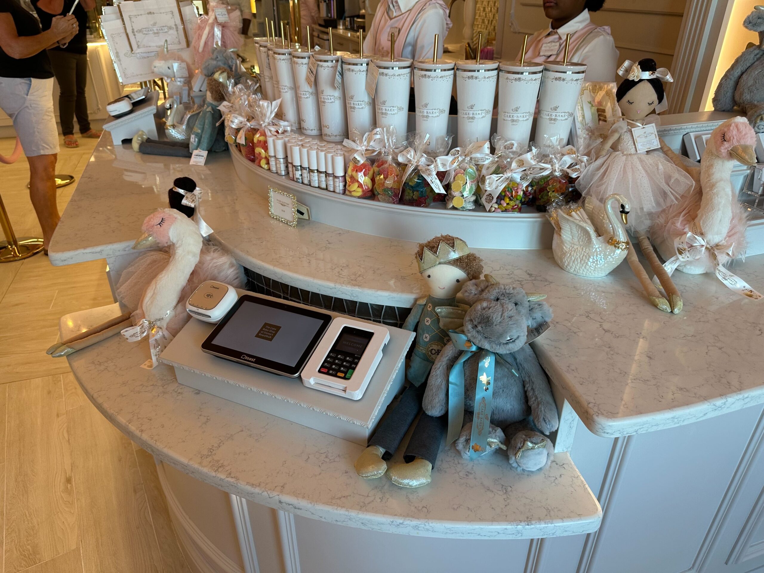 A candy store counter at Walt Disney World features plush toys, an array of candy jars, a digital payment device, and a cash register, all creating a magical shopping experience.