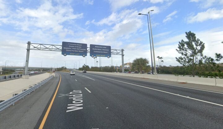 Wide highway with overhead signs for theme park directions, including Walt Disney World. Sparse traffic glides along under a blue sky, with some trees lining the side near Seven Seas Drive.