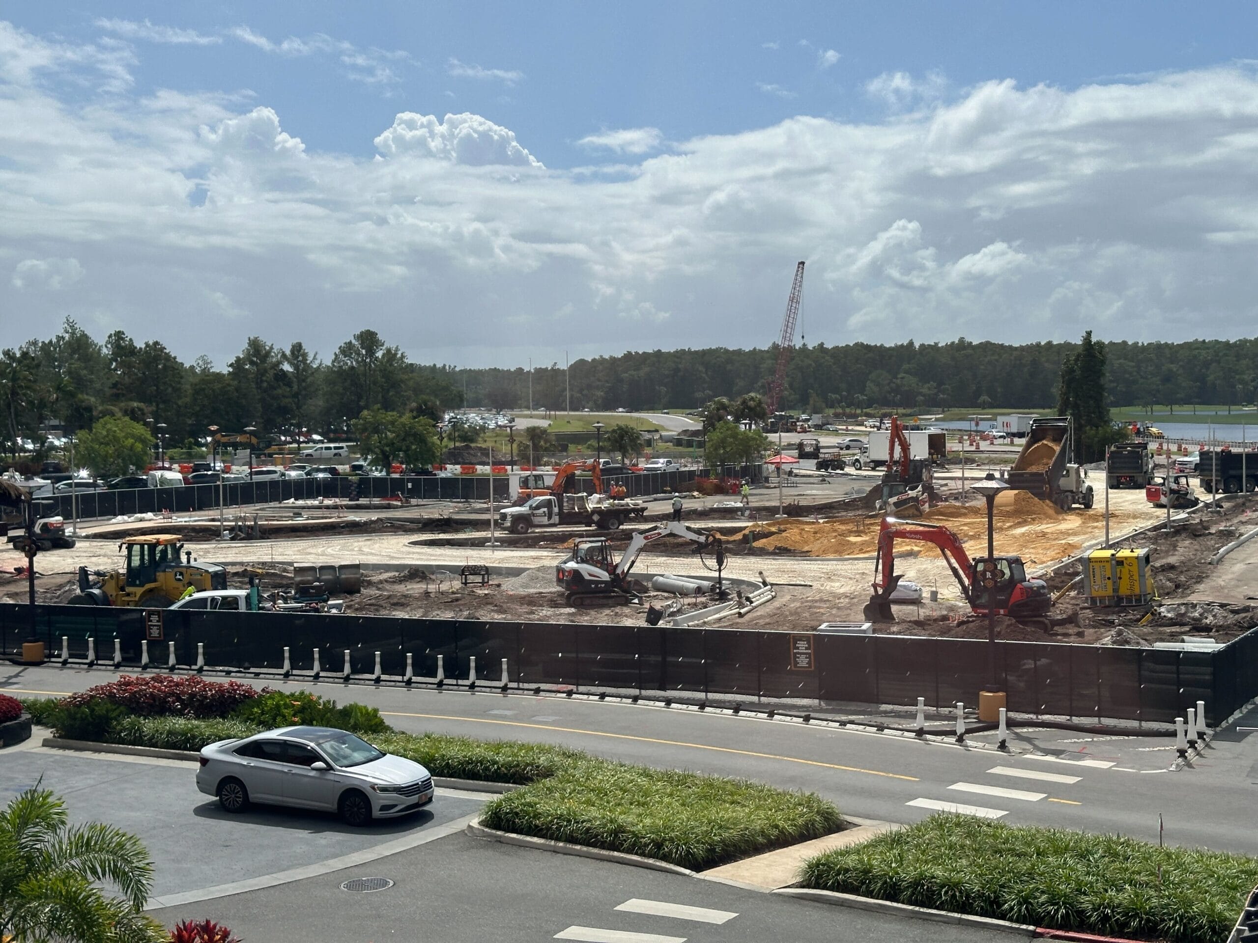 Construction site with various excavators and vehicles, bordered by fencing, trees, and a parked white car in front.