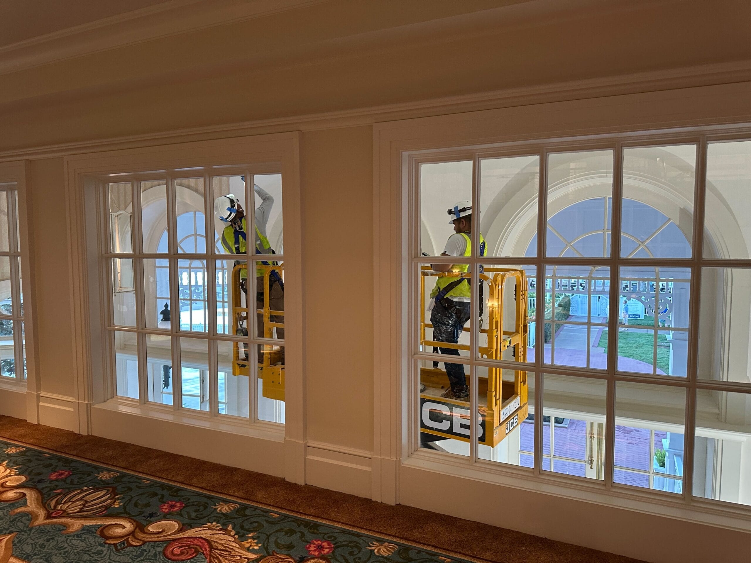 Two workers in safety vests use a lift to clean large indoor windows at the Grand Floridian overlooking an arched courtyard.