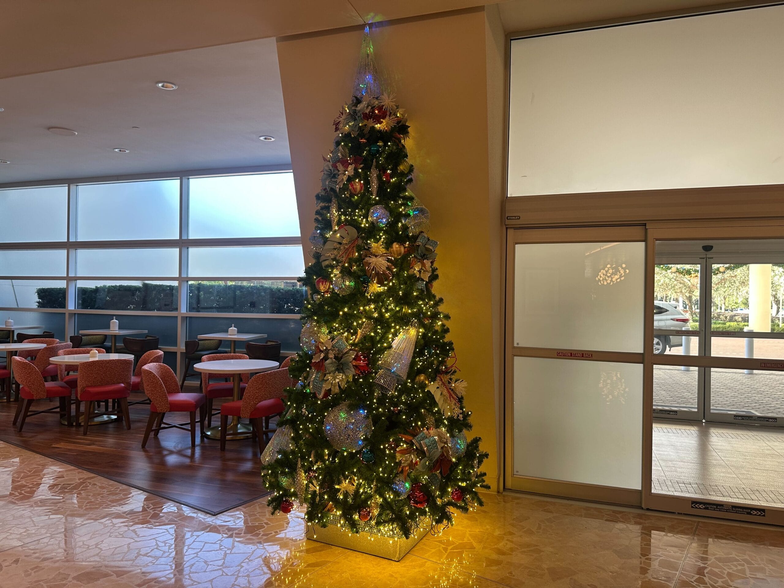 A decorated Christmas tree with lights stands in a corner of a room near tables and chairs, next to a glass door.
