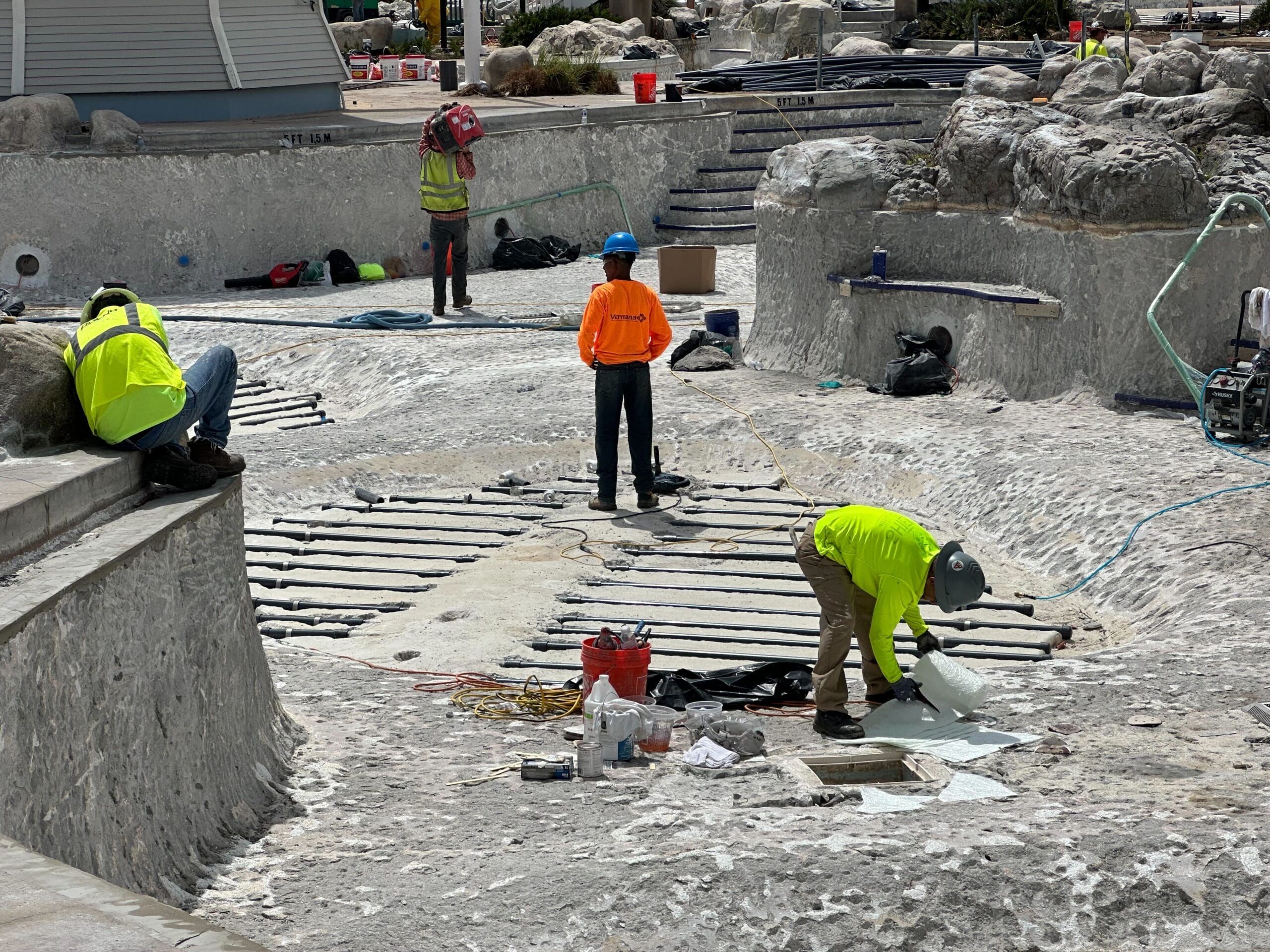Workers in bright safety clothing and helmets repair and maintain the empty, rocky outdoor pool area, with scaffolding, equipment, and tools scattered around.