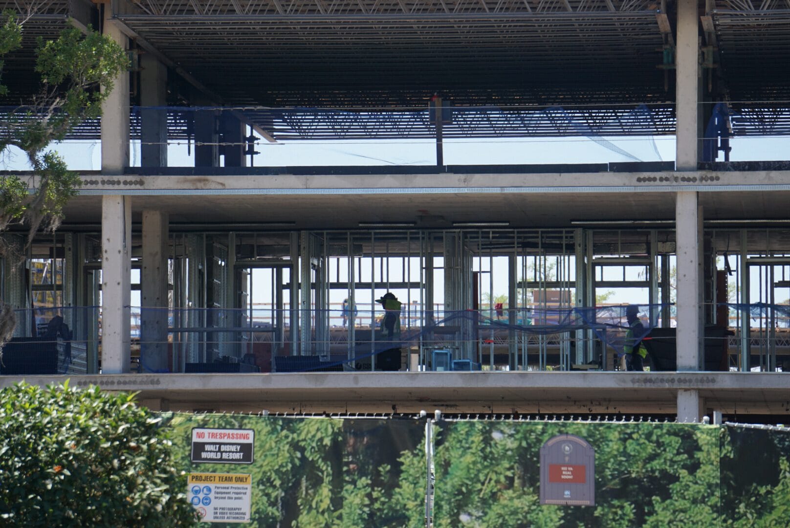 Construction site with workers inside a multi-level building structure, partially obscured by trees and a fence in the foreground.