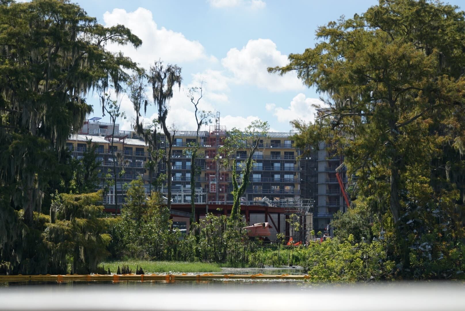 Disney Lakeshore Lodge under construction behind trees and a pond on a sunny day.