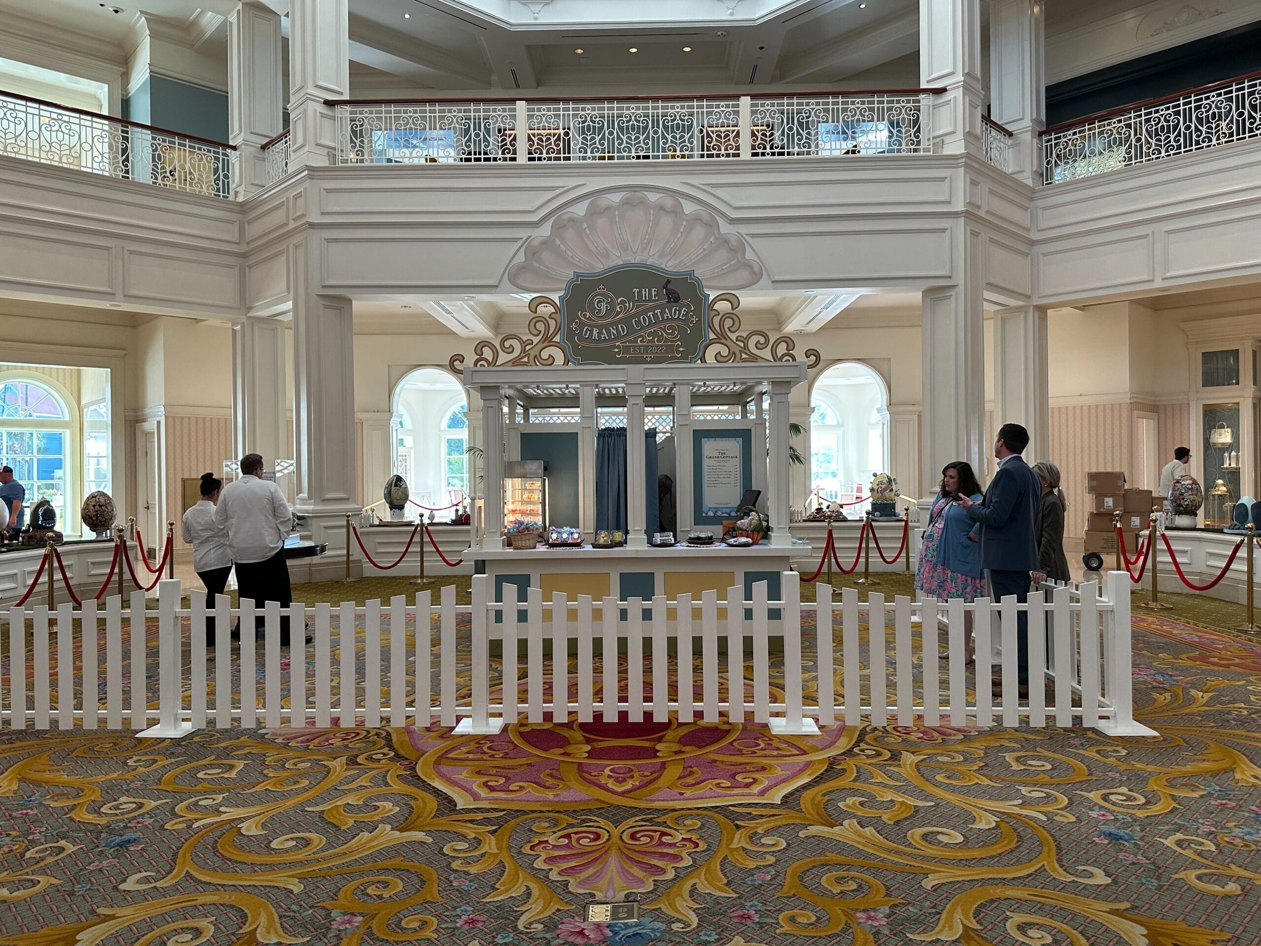A decorative booth titled The Grand Cottage is displayed in a large, elegant room with ornate railings. A white picket fence surrounds it, with people nearby and red ropes forming a queue.