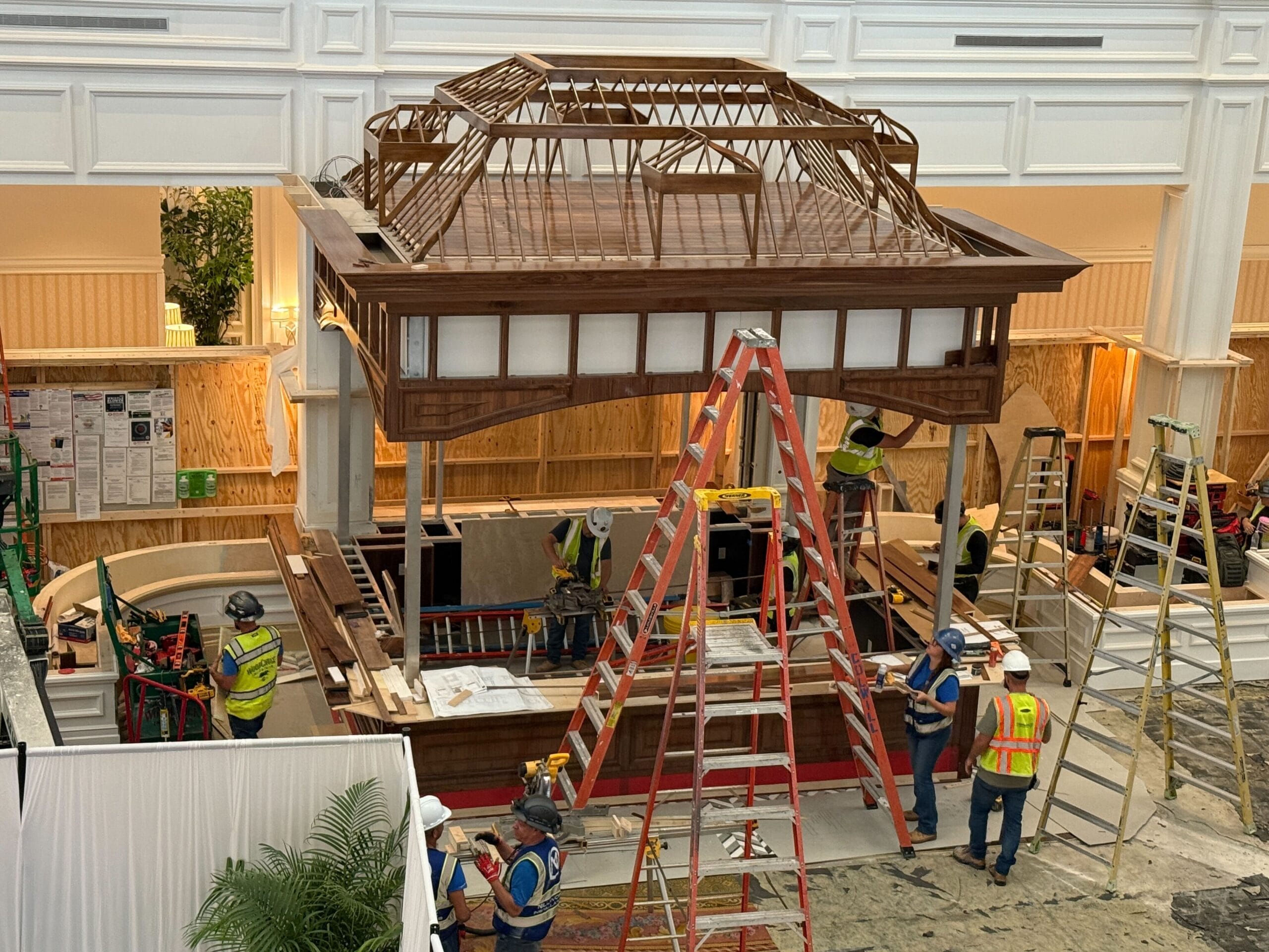 Construction workers building a wooden structure indoors, surrounded by ladders and construction materials.