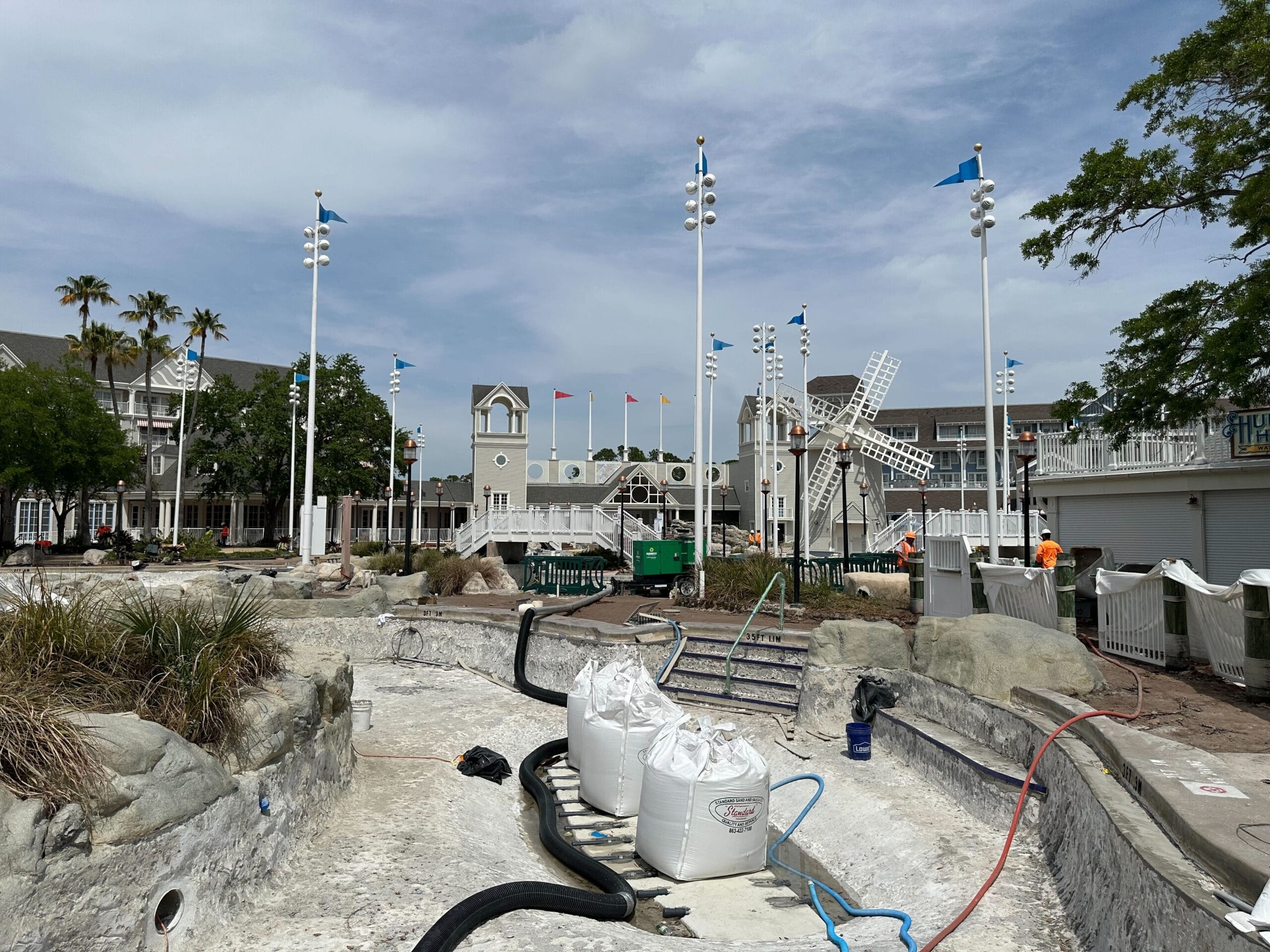 At the construction site of a drained pool area, scaffolding supports activities among rocks and equipment. Workers are busy while nearby white structures and palm trees rise under a partly cloudy sky, reminiscent of Disney's Yacht Club ambiance.