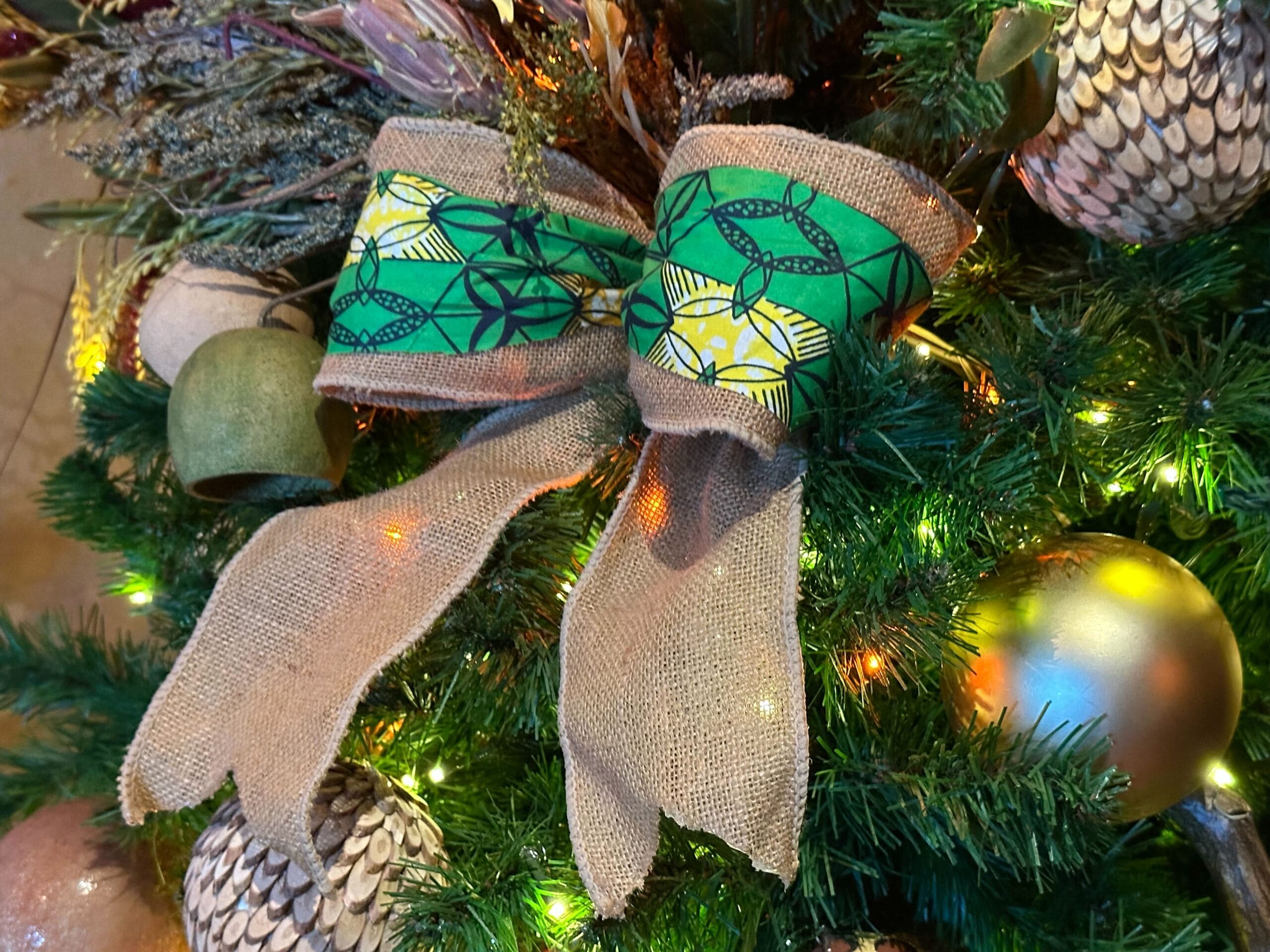 Close-up of a Christmas tree decorated with a burlap bow featuring green and yellow patterns, surrounded by colorful ornaments and lights.