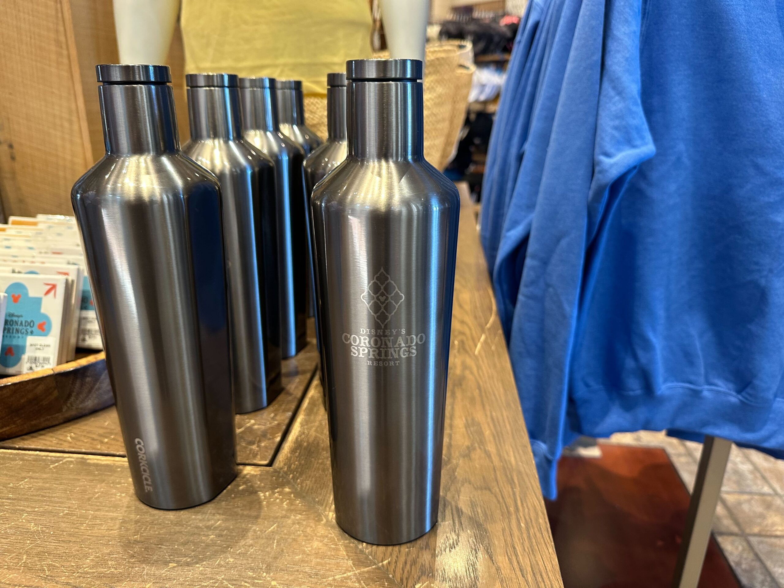 Several metal water bottles are displayed on a wooden table, showcasing the elegant Coronado Springs merchandise, with a blue shirt hanging in the background.