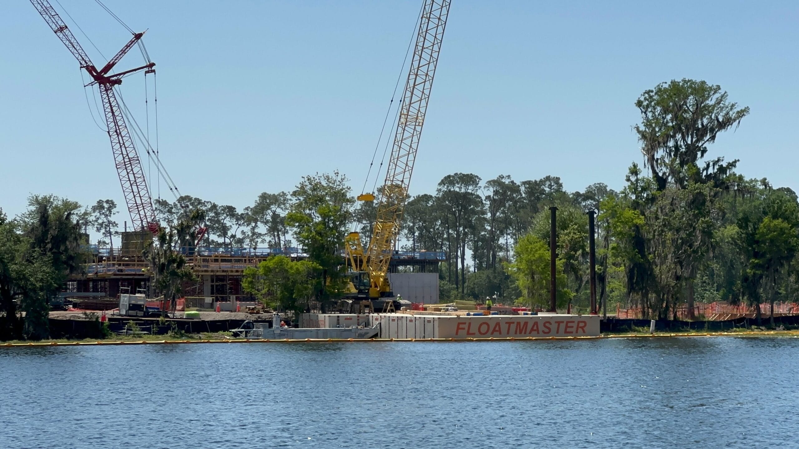 A construction site by a lake features a large crane and a barge labeled FLOATMASTER. In the background, trees sway under a clear blue sky. Stay tuned for more construction updates on this exciting Disney project near the future Lakeshore Lodge.