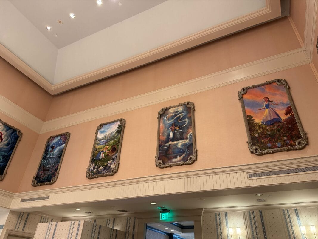 A series of framed paintings adorn the peach-colored wall in a gallery-like setting, reminiscent of Park Fare Art at the Grand Floridian Resort, all beautifully illuminated by recessed lighting.