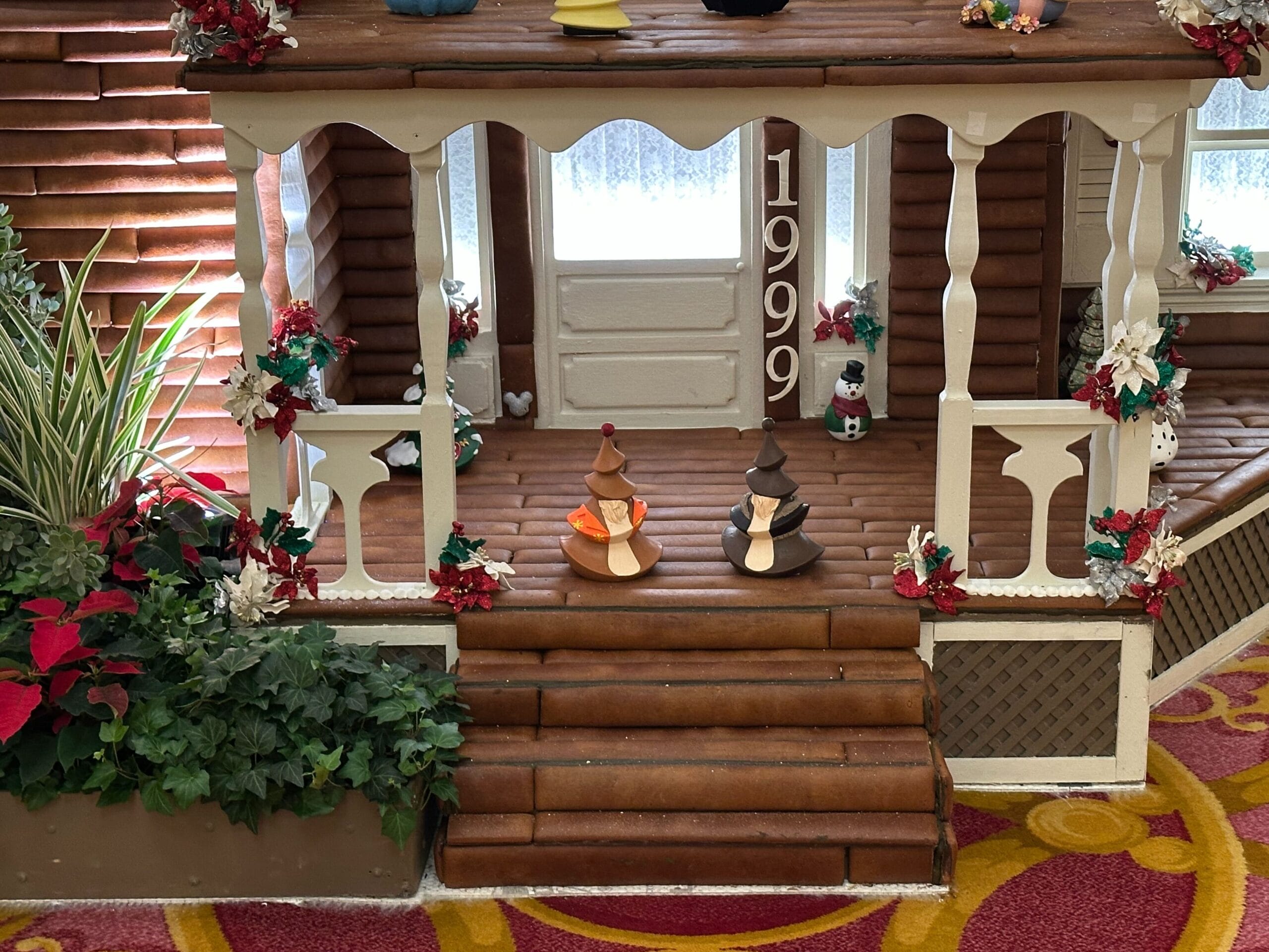 A miniature porch scene with candy figures on the steps, the year 1999 displayed on a post, and festive decorations with plants and flowers around.