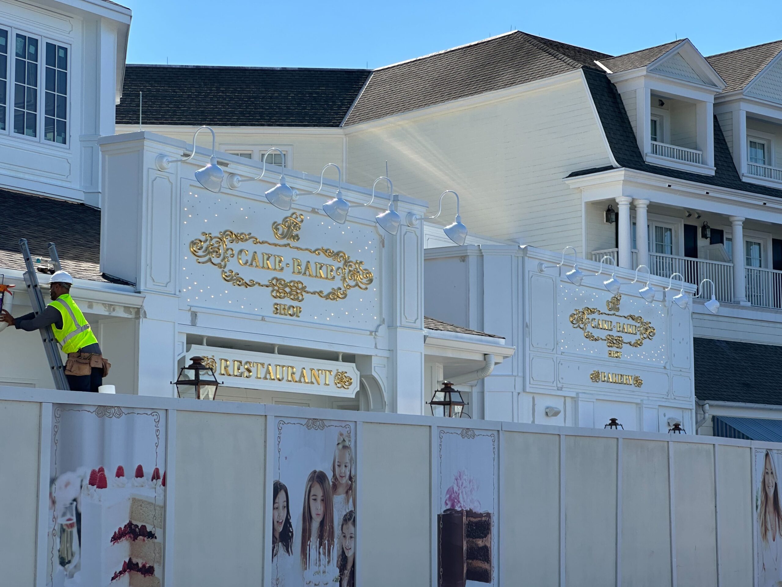 A person in a yellow vest stands on a ladder next to a white building with ornate signage that reads Cake Bake Shop and Restaurant. A construction barrier features images of desserts and people.