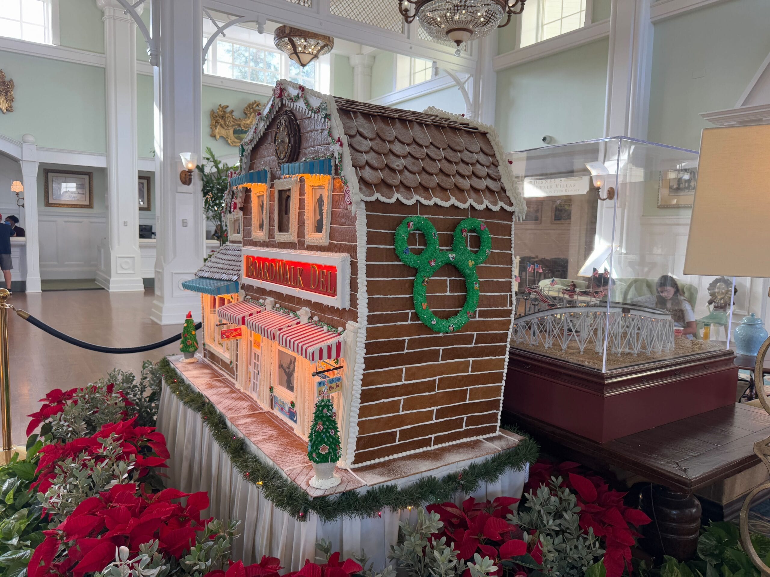 Large gingerbread house display with decorative lights, a green wreath, and candy accents. Surrounded by red poinsettias inside a bright room.
