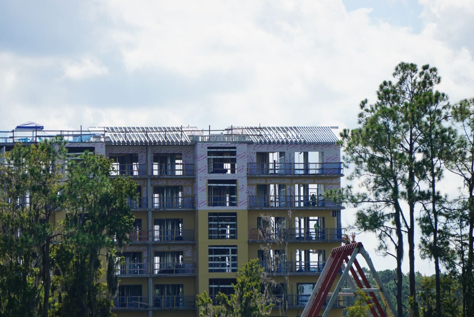 A multi-story Disney Lakeshore Lodge under construction, partially obscured by trees, with a cloudy sky in the background.