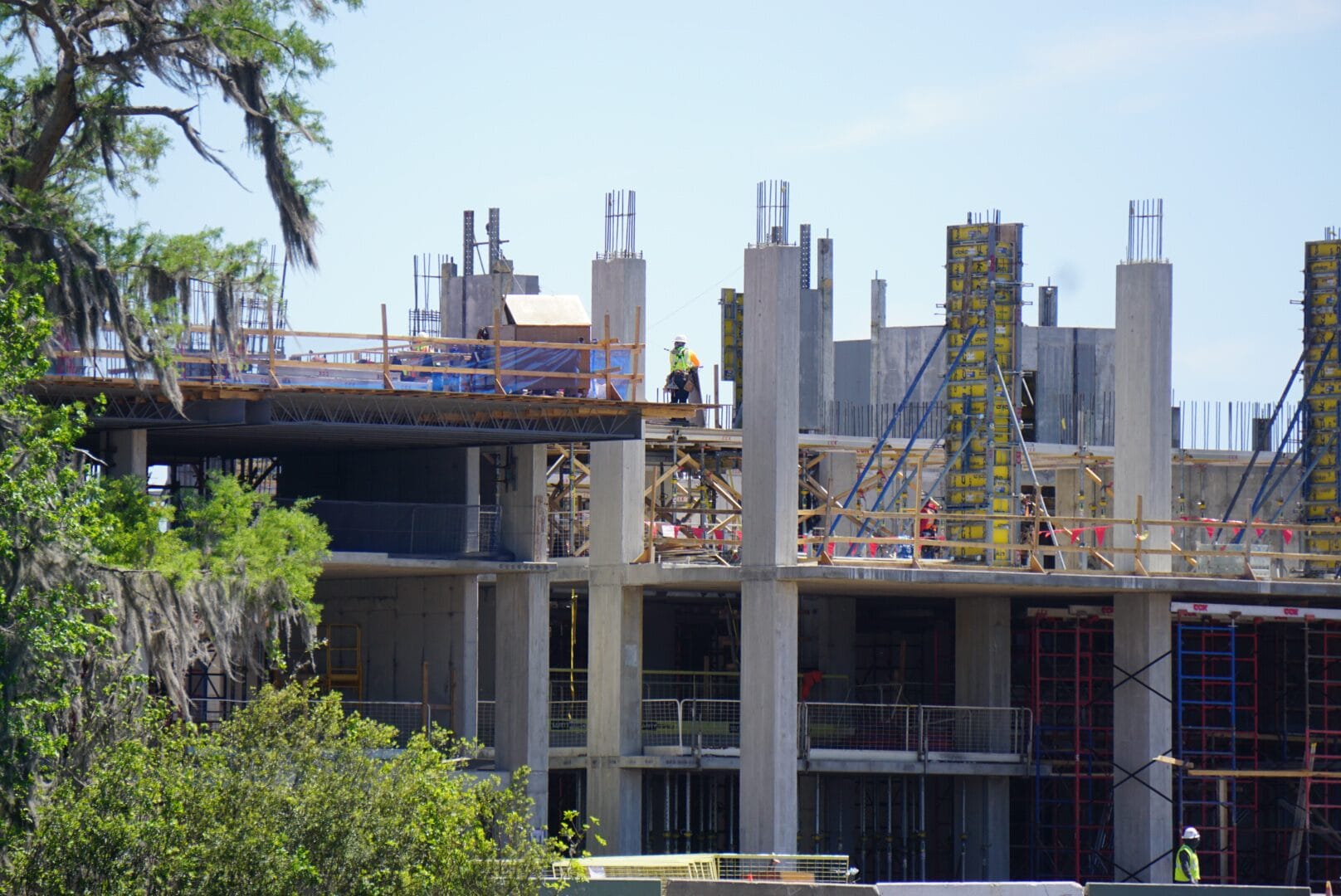 Construction site with partially completed multi-story building. Workers and scaffolding are visible. Trees and a clear sky are in the background.