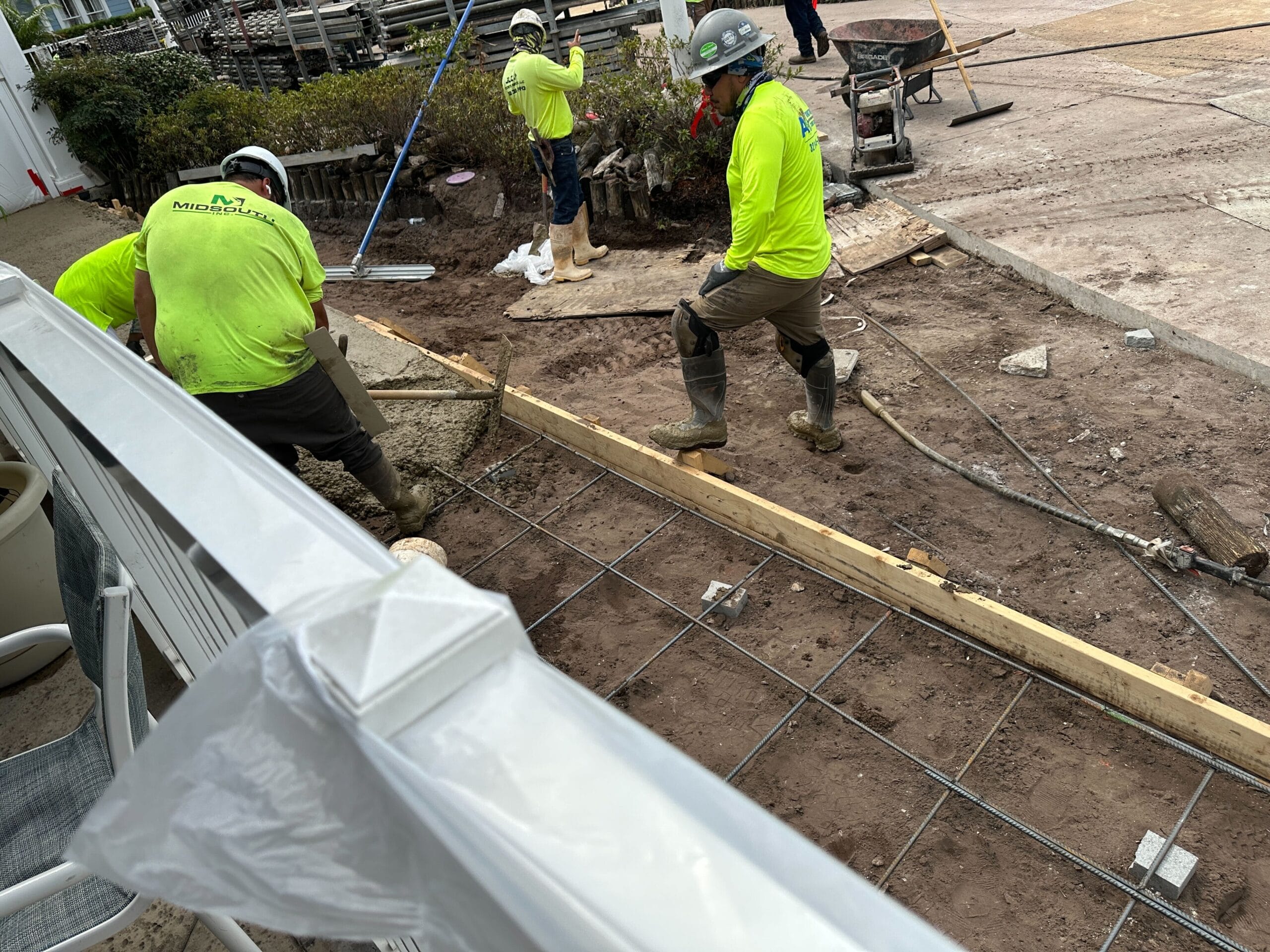 Construction workers in yellow shirts pour and level concrete on a rebar-framed sidewalk under construction.