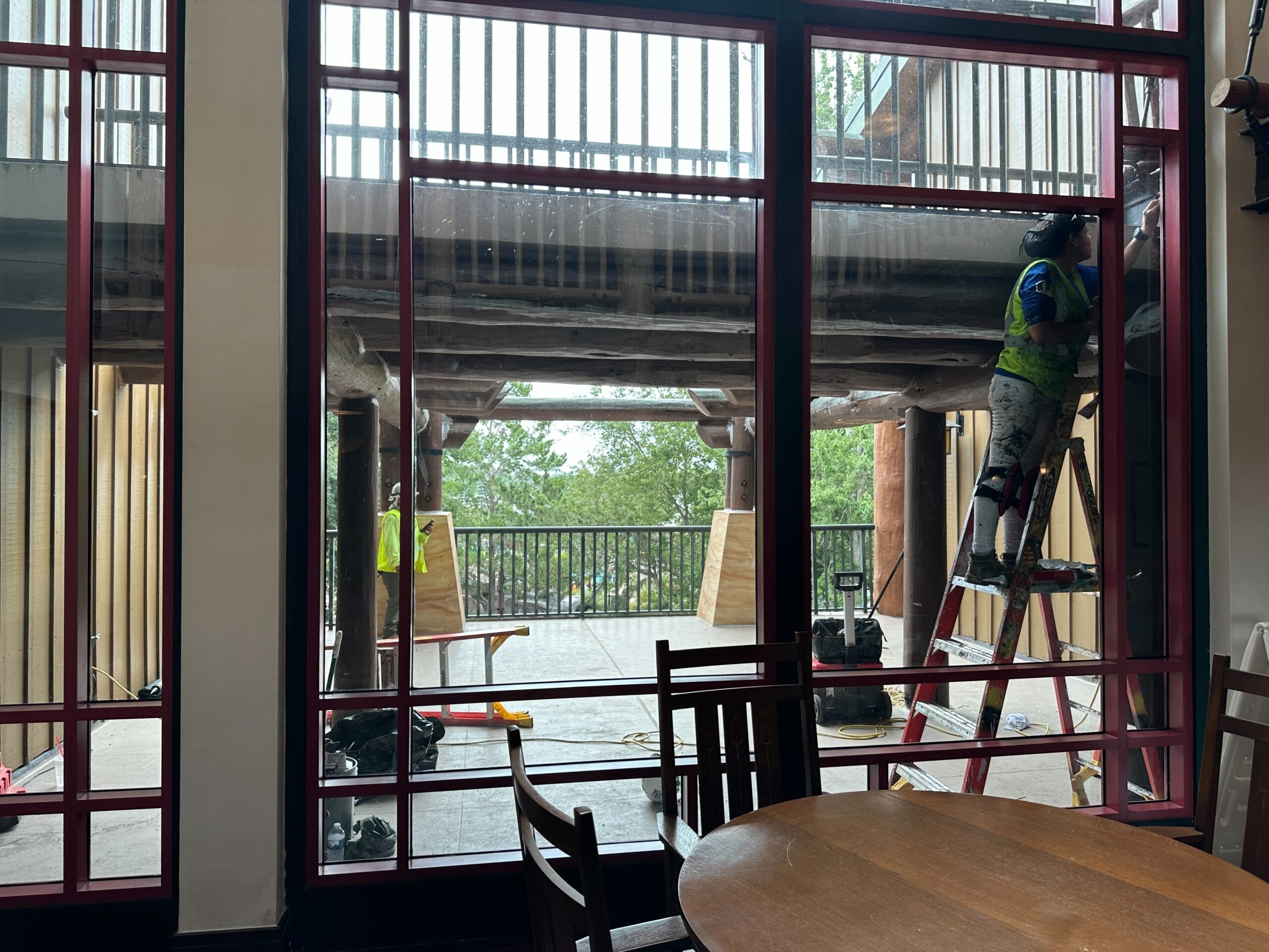 Worker on a ladder repairs a wooden balcony at Wilderness Lodge, viewed through large glass doors from indoors.