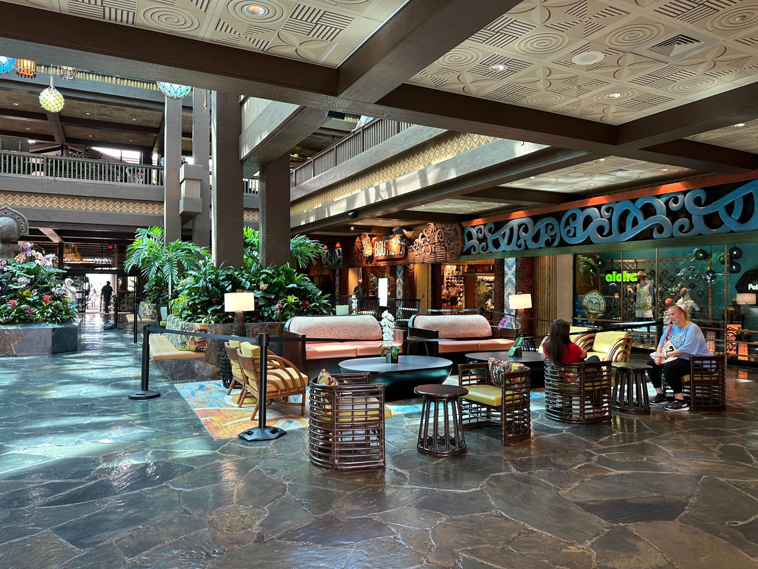 Spacious hotel lobby with tropical plants, modern seating, and people relaxing near colorful decor and large windows.