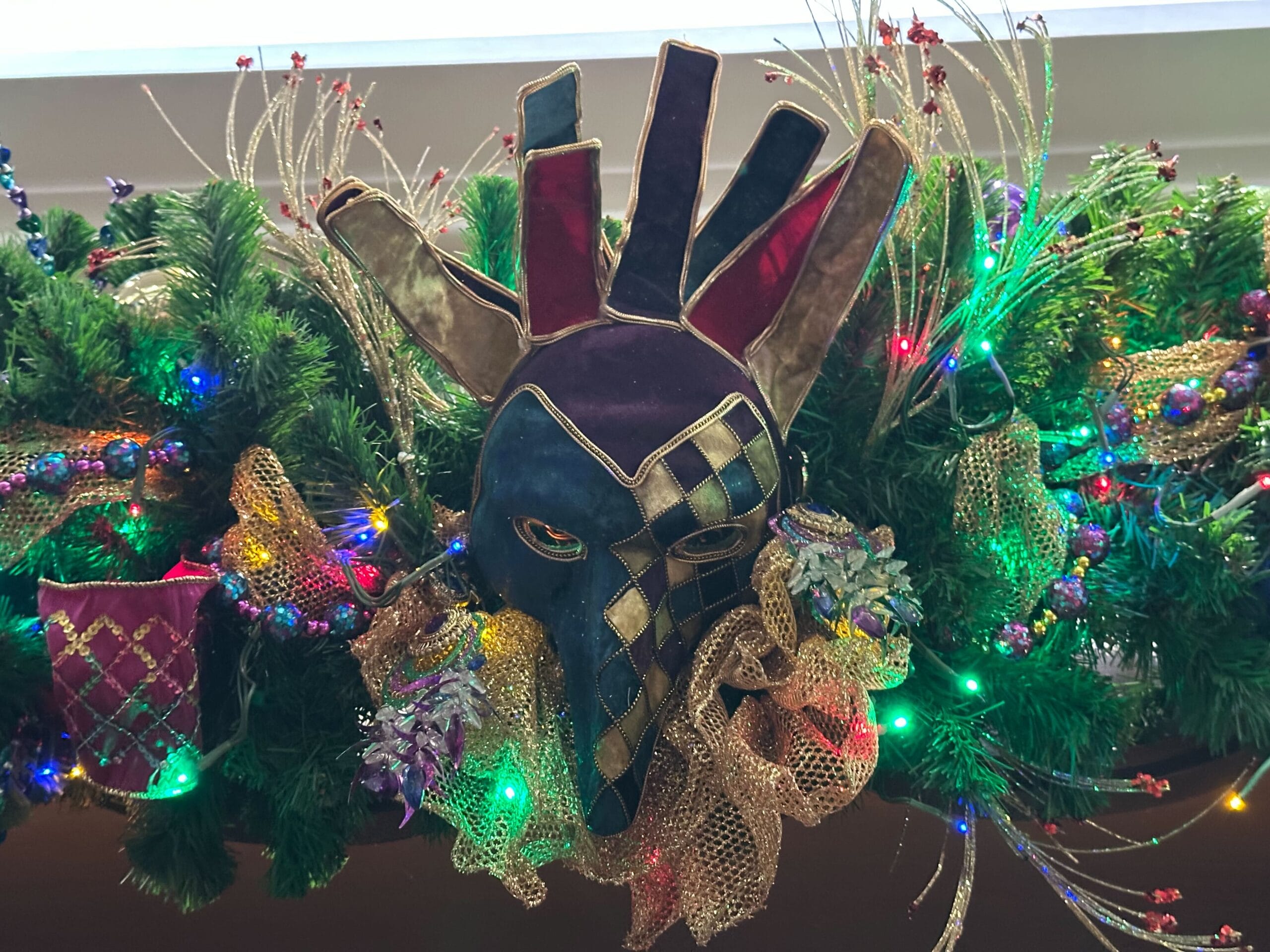 Festive jester mask with multicolored sections and diamond patterns, surrounded by greenery, lights, and decorative ribbons.