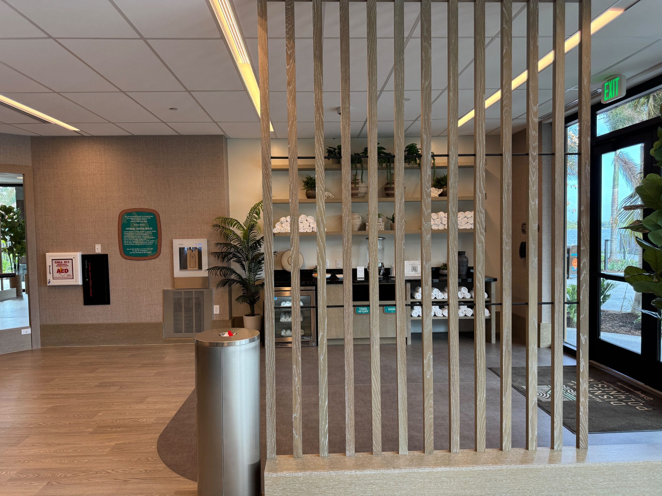 A modern hotel lobby with a wooden divider, potted plants, and a refreshment area with coffee supplies. Large windows provide natural light.