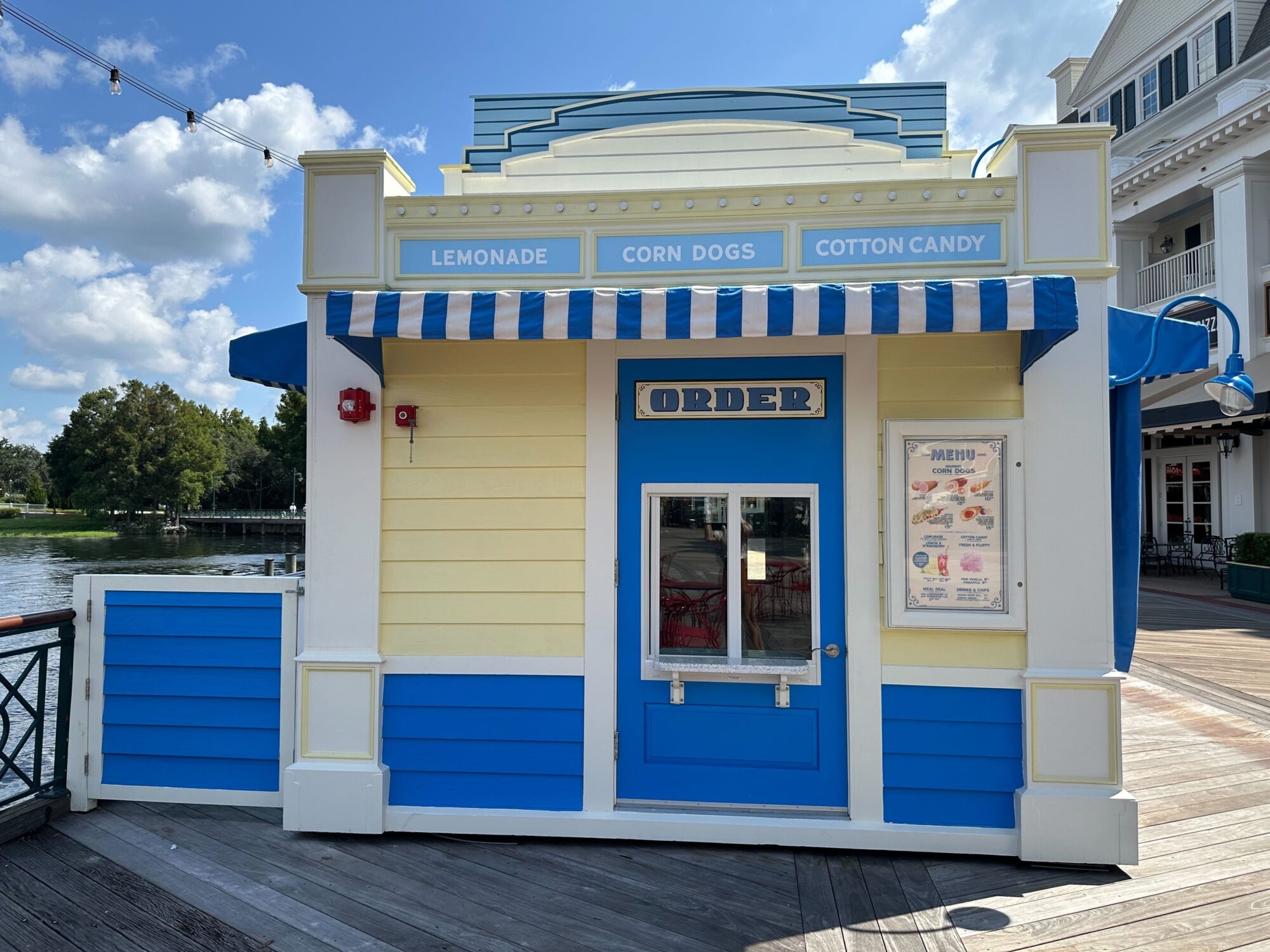 A small yellow and blue food stand by the water with a sign advertising lemonade, corn dogs, and cotton candy. A menu is posted next to a blue door labeled Order.