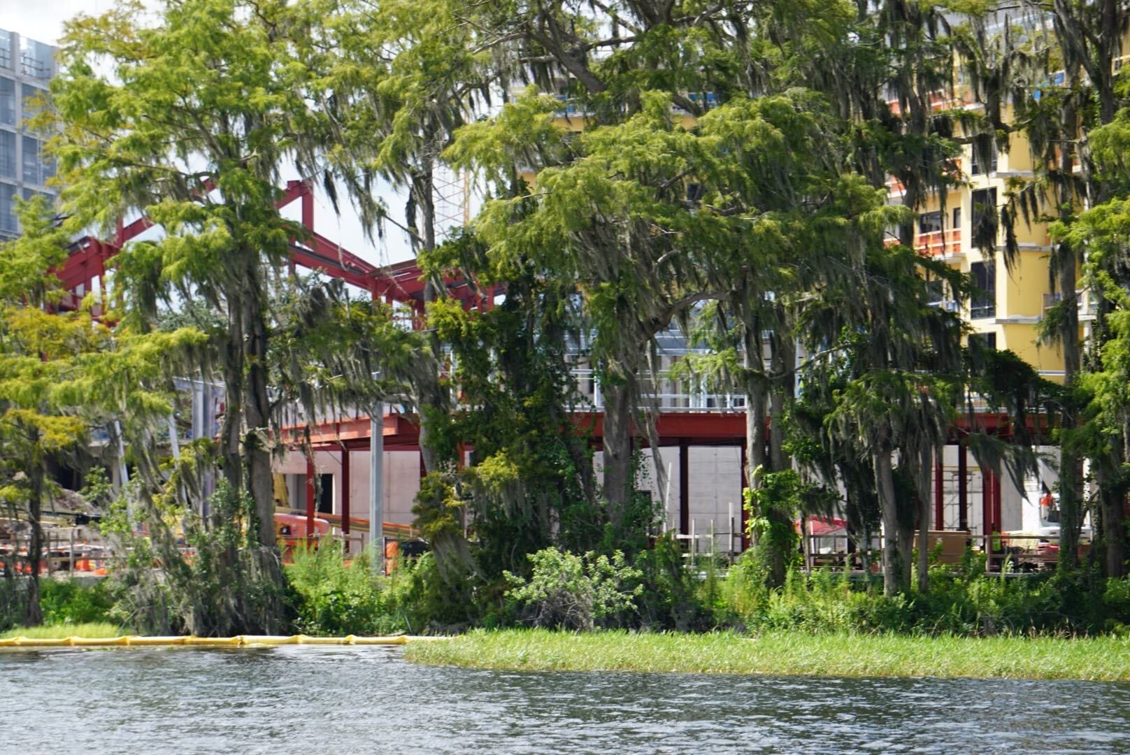 Trees with hanging moss partially block the view of a building under construction by the water's edge.