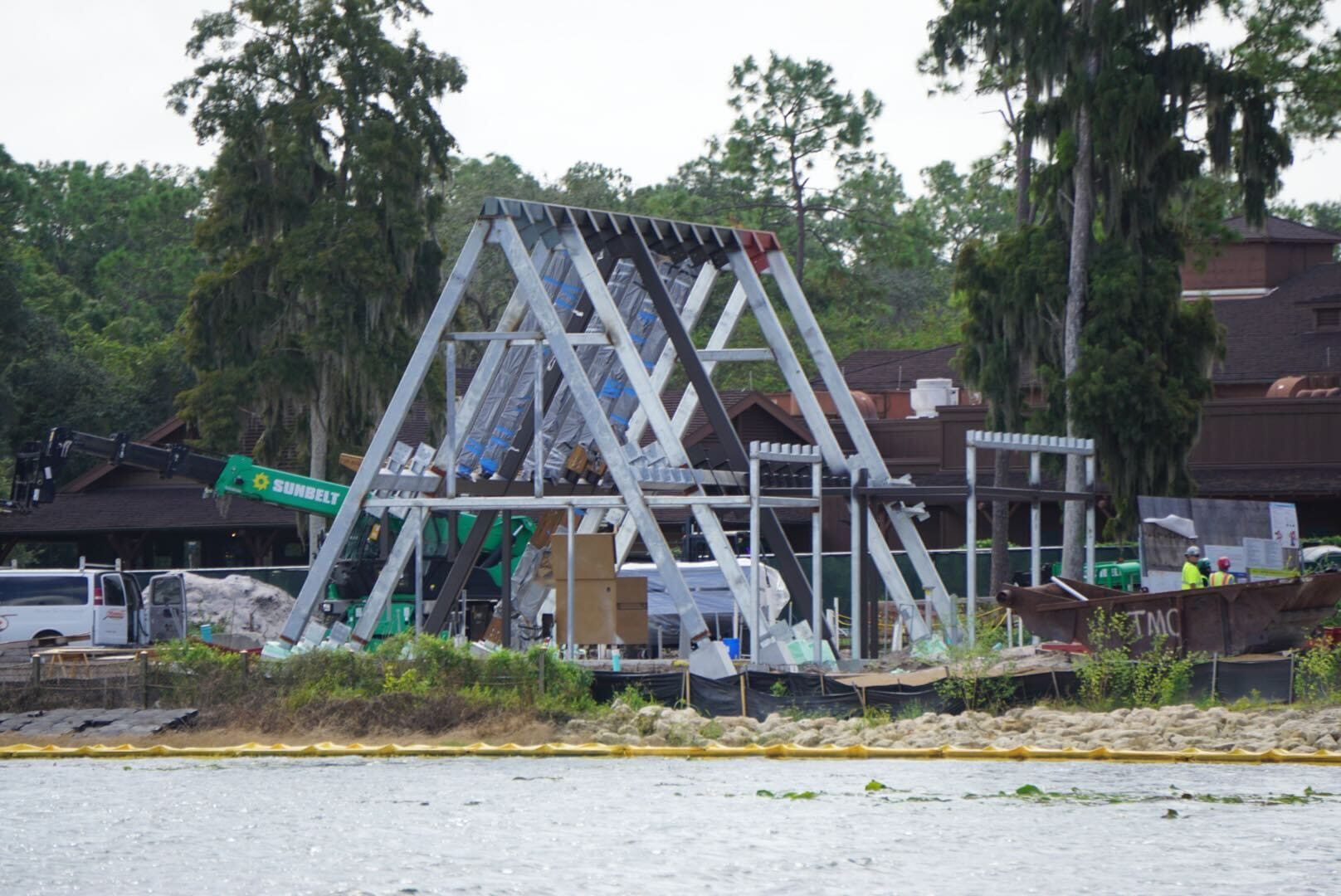 Steel frame structure under construction near water at Lakeshore Lodge, with vehicles and workers among trees.