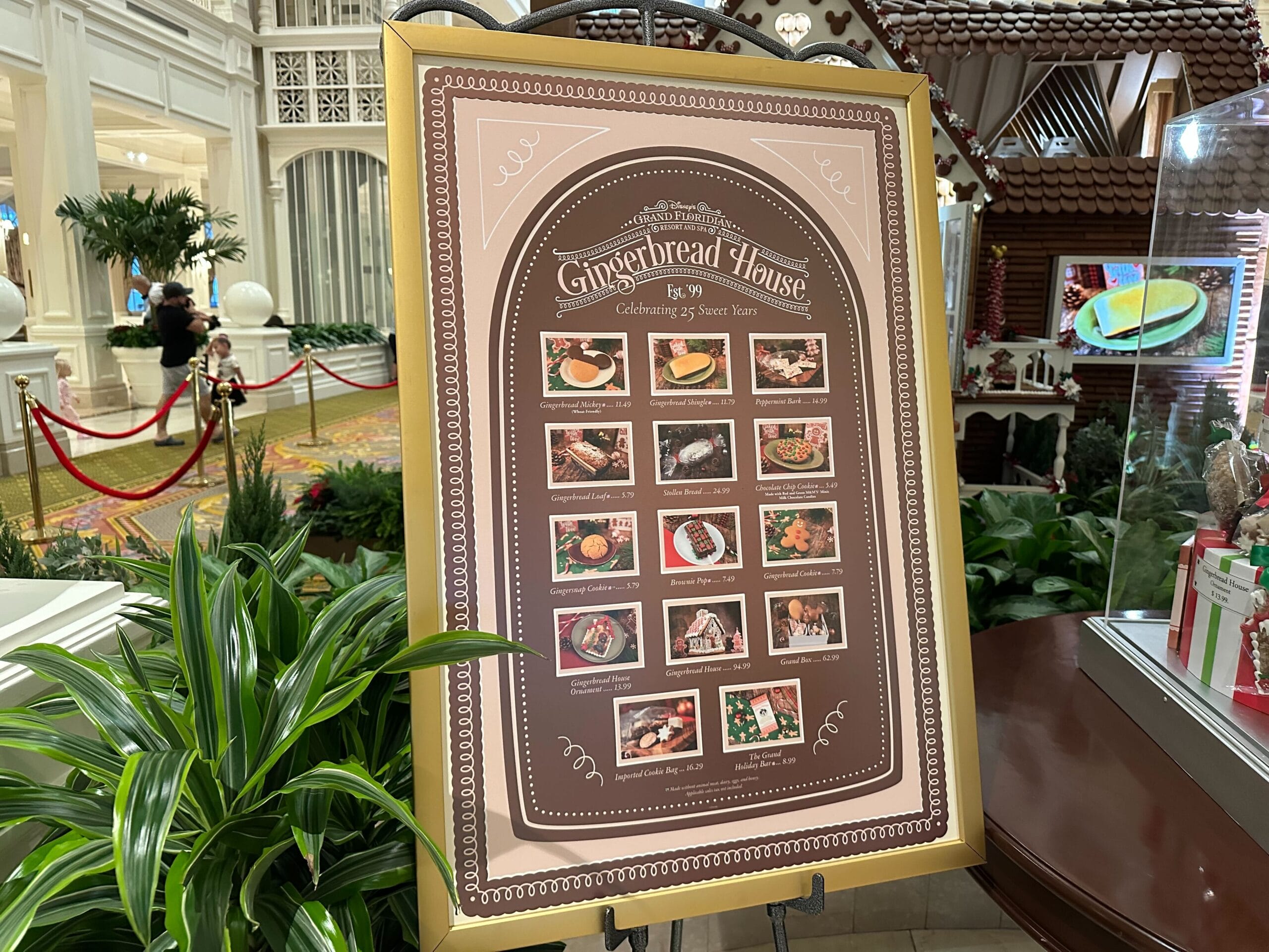 A decorative menu board at the Grand Floridian Gingerbread House display showcases various holiday-themed food items and desserts, set amidst a festive indoor setting.