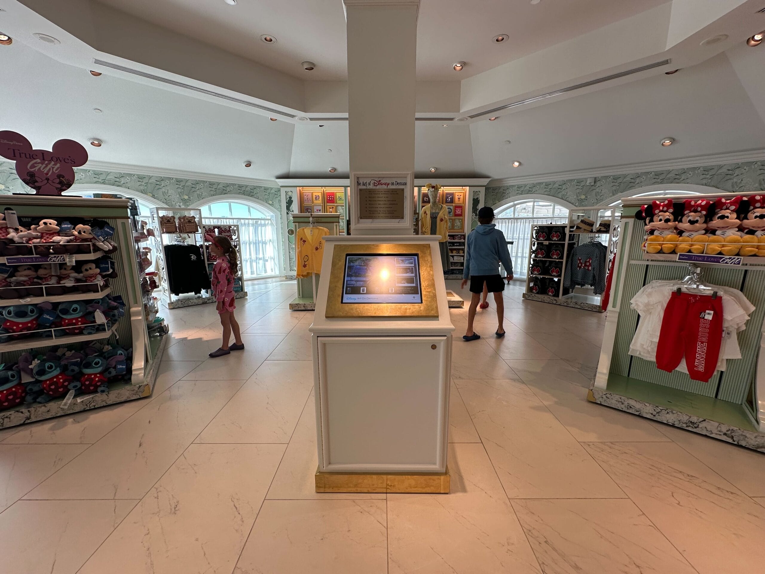 A well-lit Disney-themed store in the Grand Floridian boasts display shelves filled with plush toys, shirts, and unique merchandise. An on-demand kiosk takes center stage, allowing guests to explore custom options while a few customers browse in the background like living art.