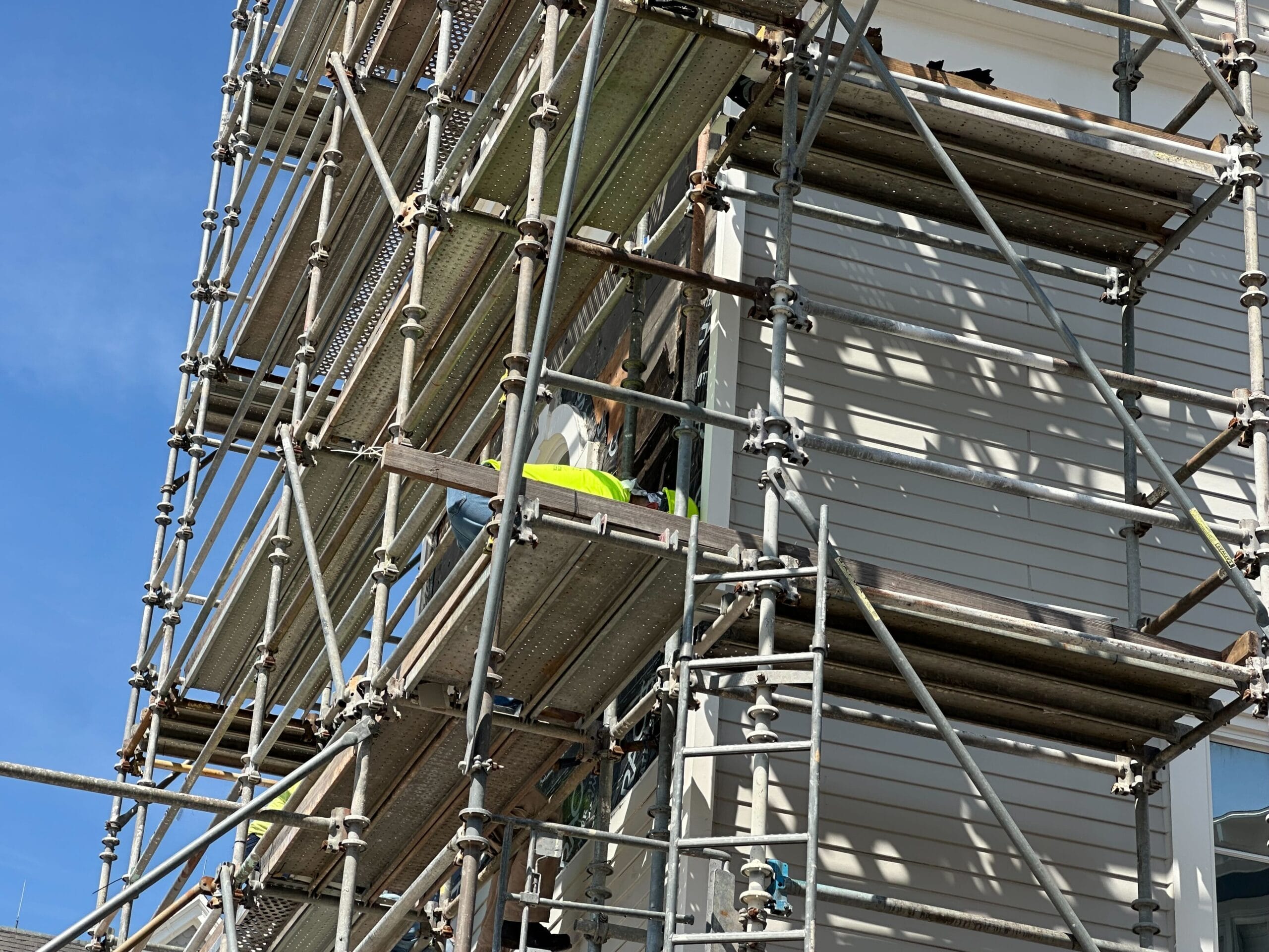 Scaffolding surrounds the exterior like a tower of new scaffolding. A worker in a bright yellow vest is visible within the structure, diligently attending to tasks. Above, the sky is clear and blue, painting an idyllic backdrop reminiscent of Stormalong Bay’s tranquility.