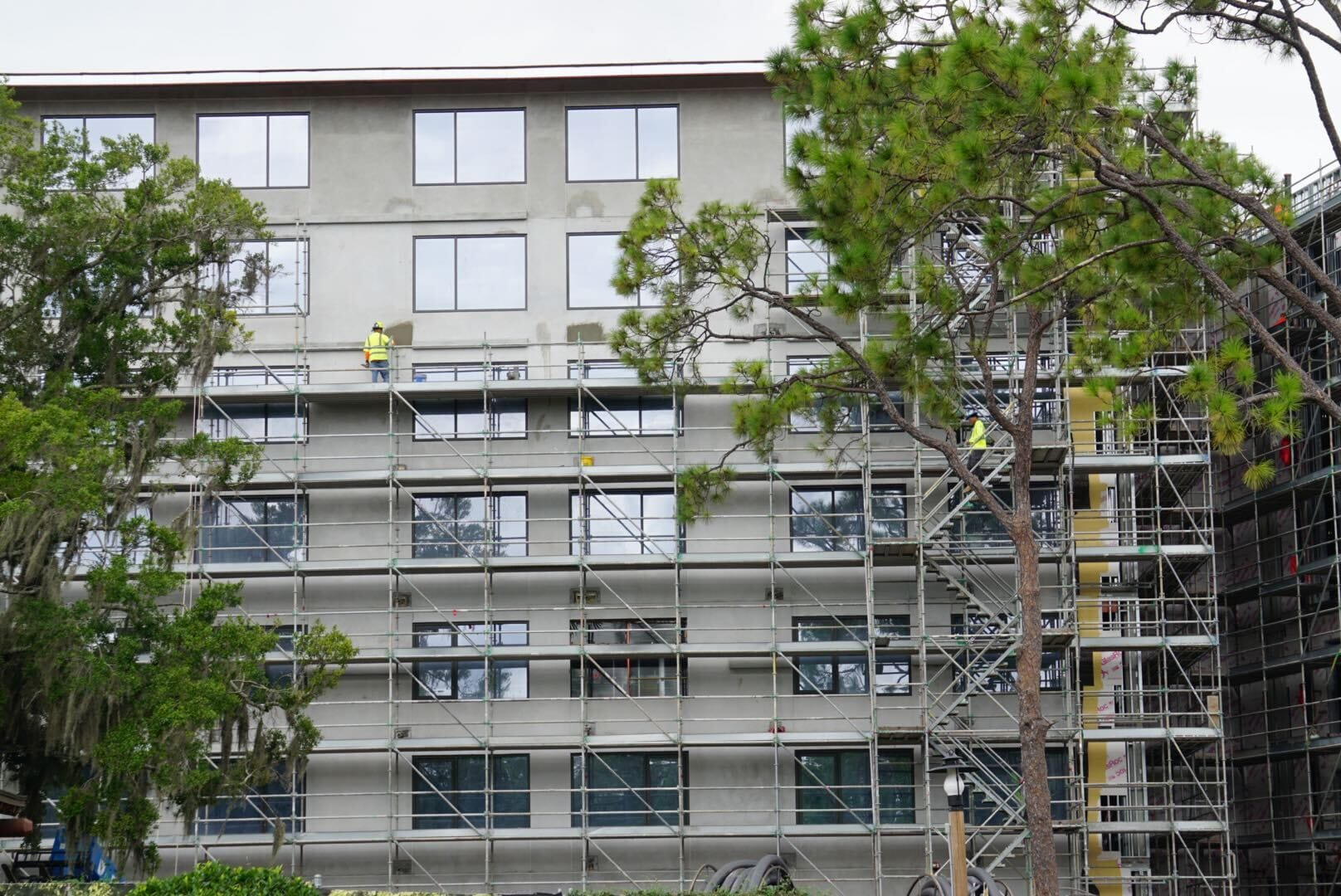Several construction workers on scaffolding update the exterior of a Lakeshore multi-story building surrounded by trees.