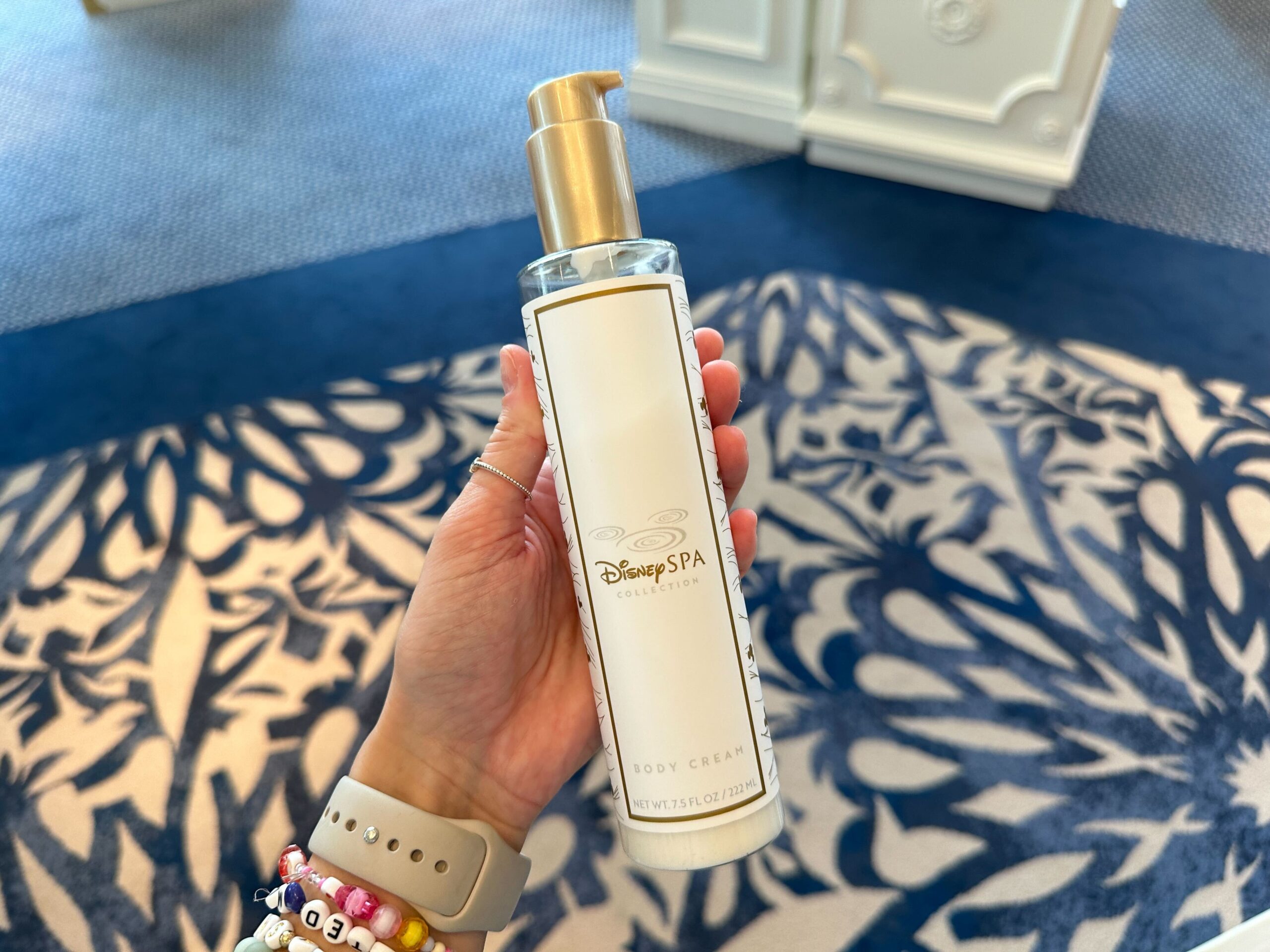 A hand holds a Disney Spa Collection body cream dispenser, now part of the Grand Floridian's enchanting new merchandise, against a patterned background.