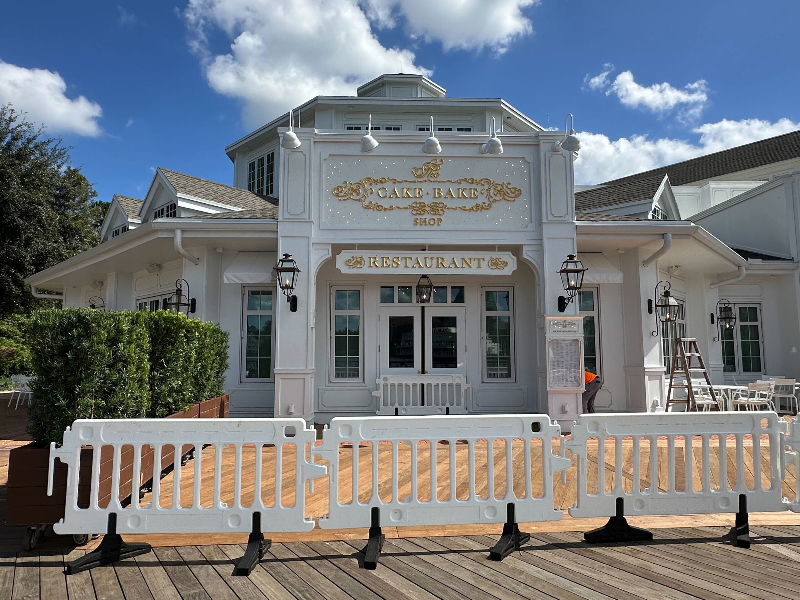 The front view of the Cake Bake Shop features elegant gold lettering, surrounded by temporary white barricades, while new seating has been added. A menu is posted near the entrance under the clear skies at Disney's BoardWalk.