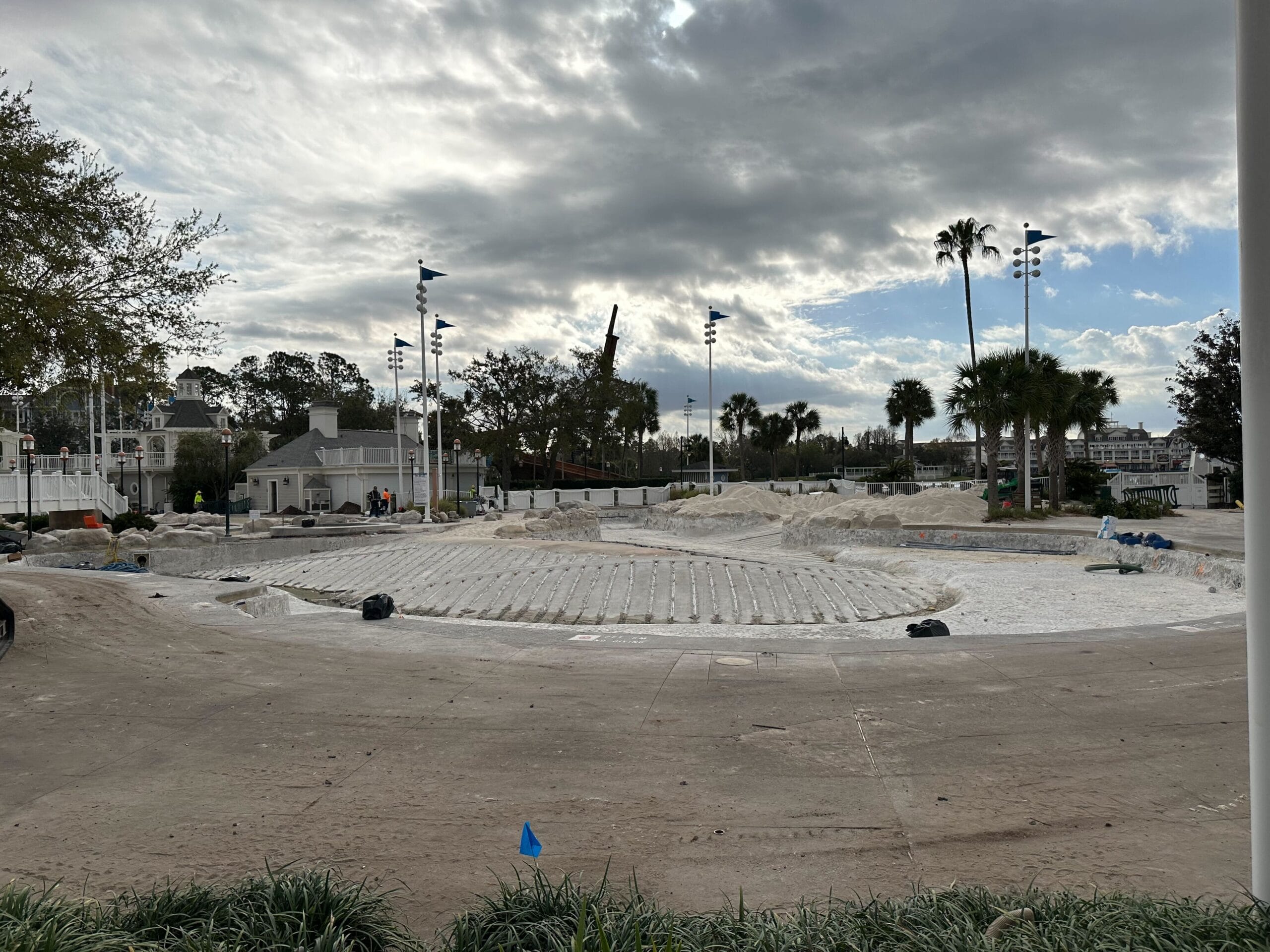 A sandy construction site is shown with palm trees and a cloudy sky. Workers and equipment are present under a partly cloudy sky.