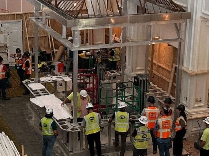 Construction workers renovating an enclosed area inside a large, ornate hotel lobby with chandeliers and patterned carpet.