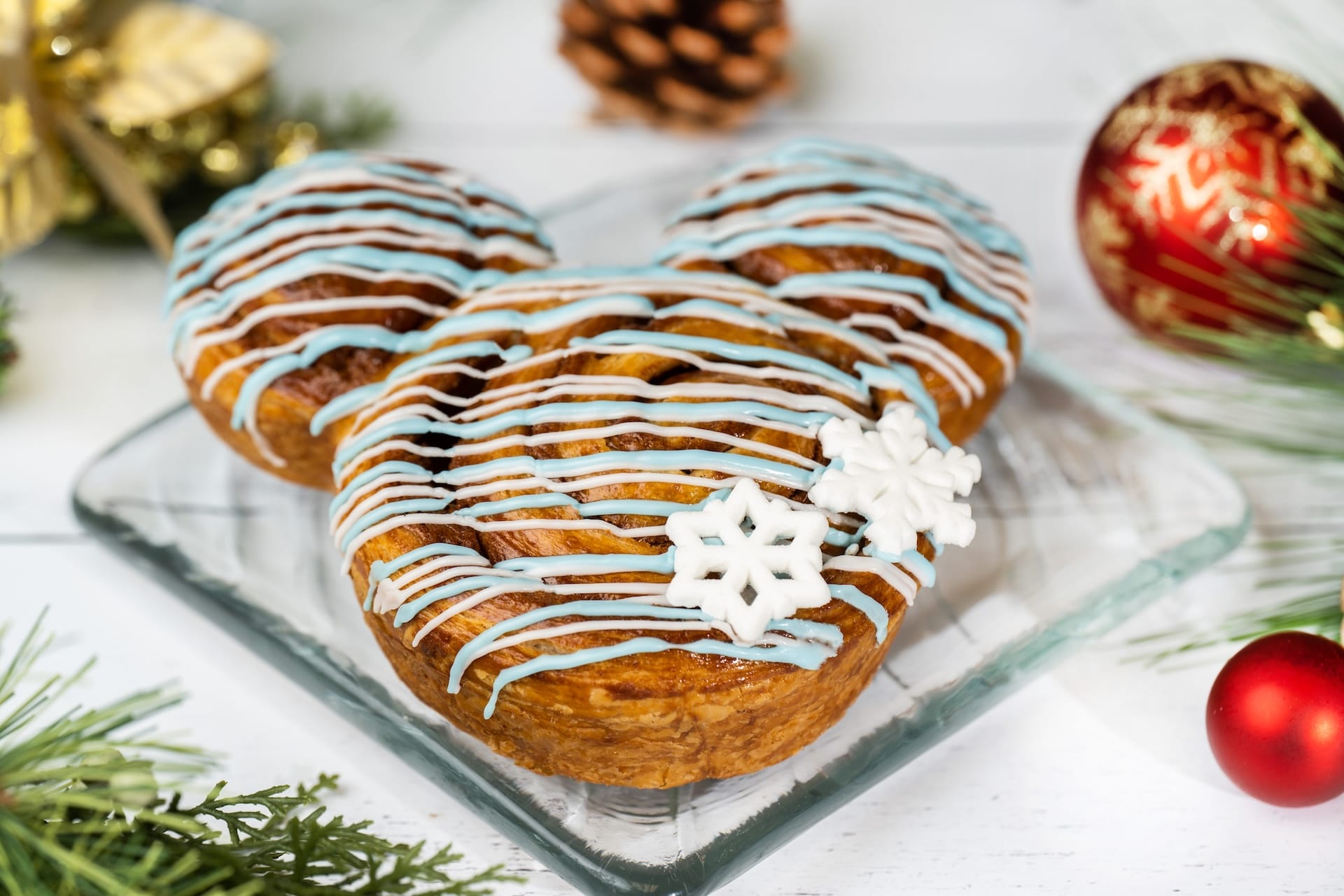 A Mickey Mouse-shaped pastry with blue and white icing and snowflake sprinkles, festive Disney holiday decor in the background.