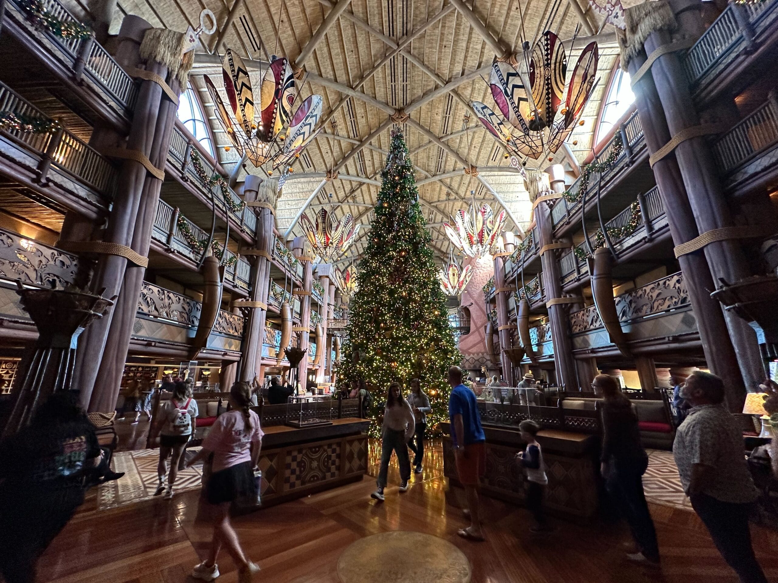 A large decorated Christmas tree stands in the center of a grand, ornately designed atrium with high ceilings and intricate lighting fixtures. People walk around the spacious area.