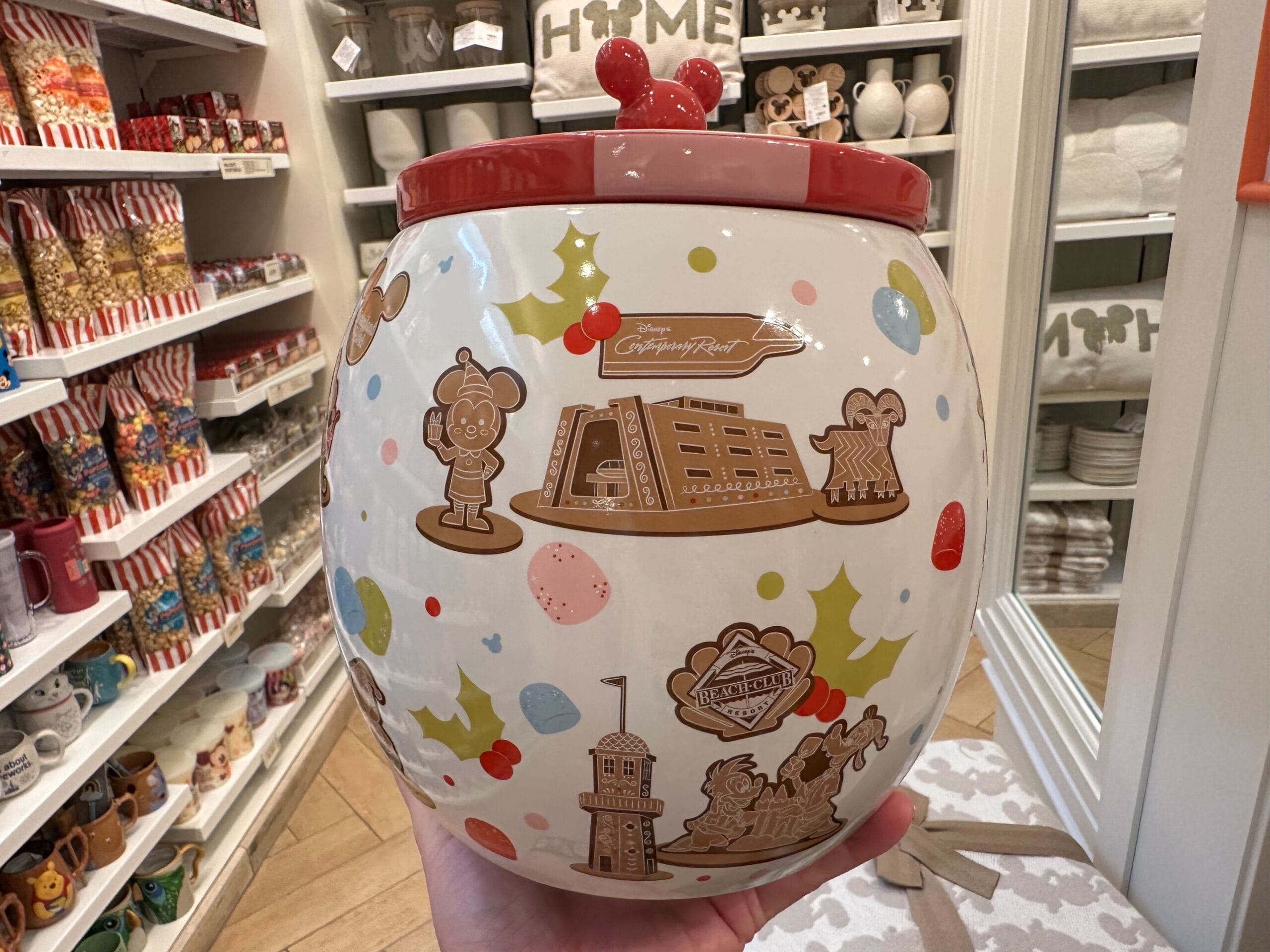 A hand holds a festive cookie jar adorned with holiday-themed Disney images, featuring Mickey Mouse and the iconic Grand Floridian Gingerbread House, inside a store brimming with similar merchandise.