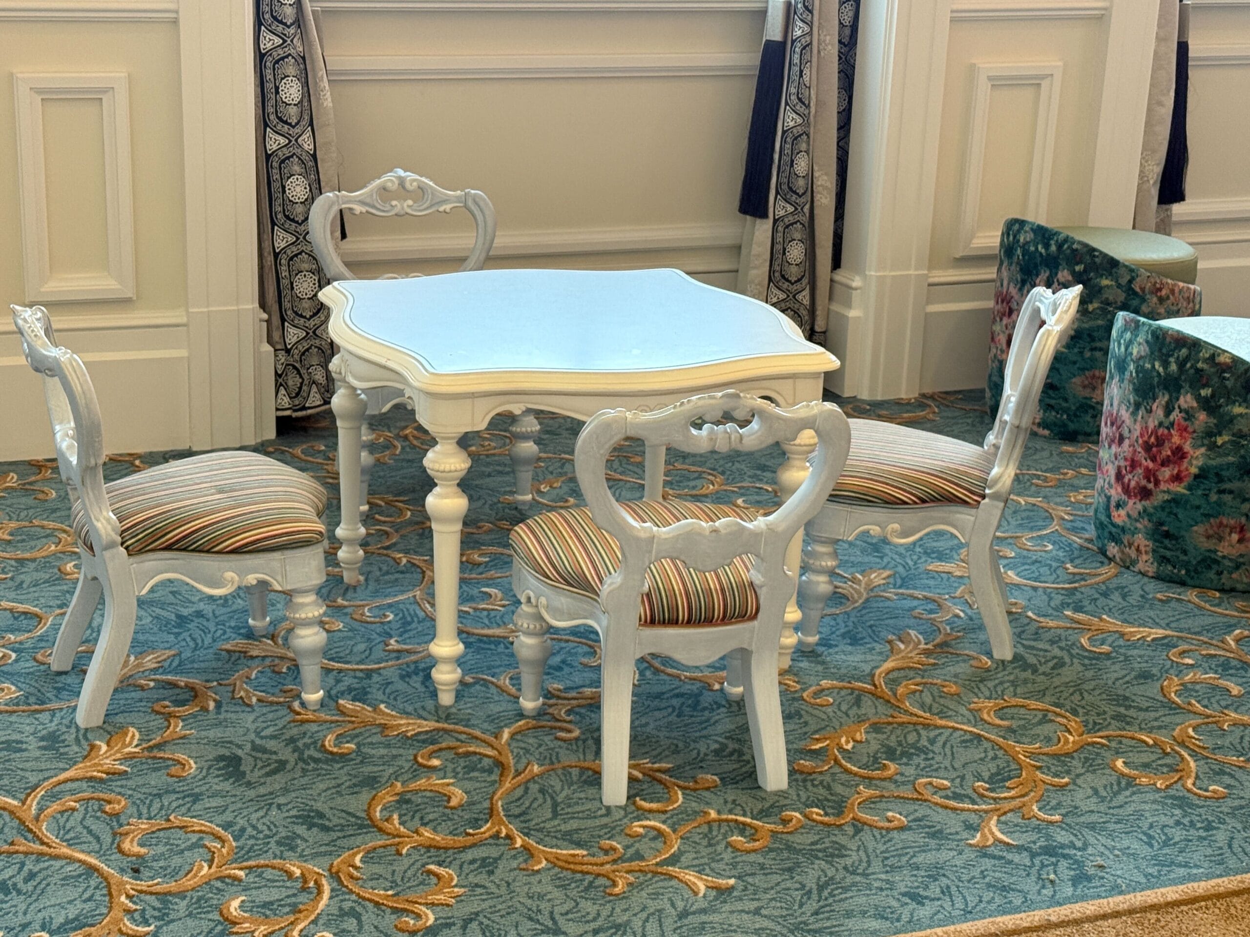 White vintage table with three matching chairs on a blue floral carpet; ottomans in the background.