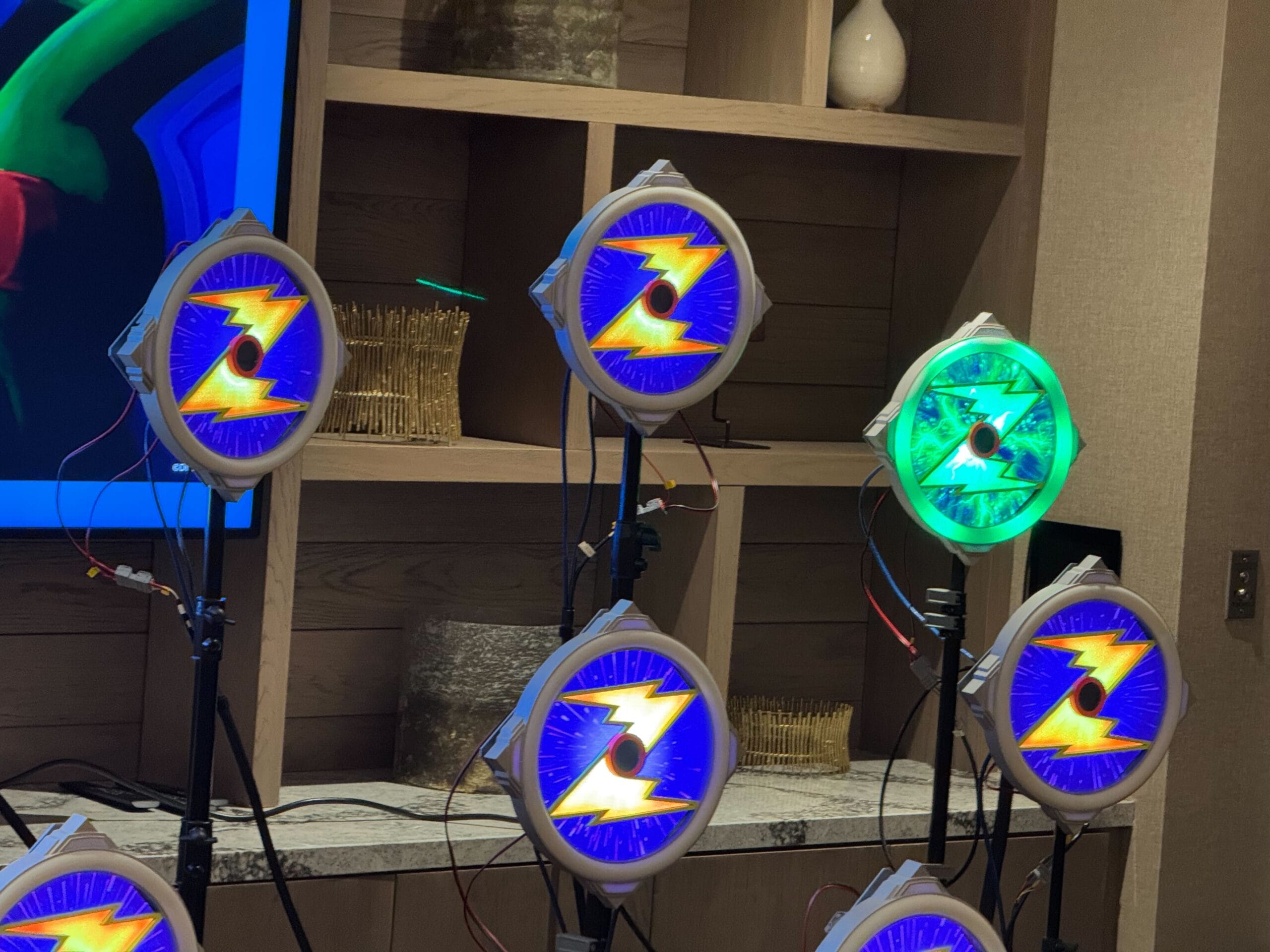 Multiple circular lights on stands with blue and yellow lightning bolt designs, one glowing green. They are set up in a room with wooden shelves in the background.