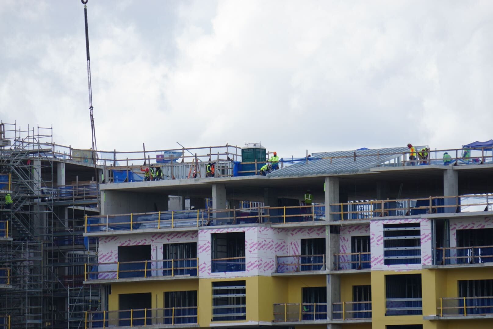 Construction workers wearing safety gear work on the roof and scaffolding of a multi-story building under construction.