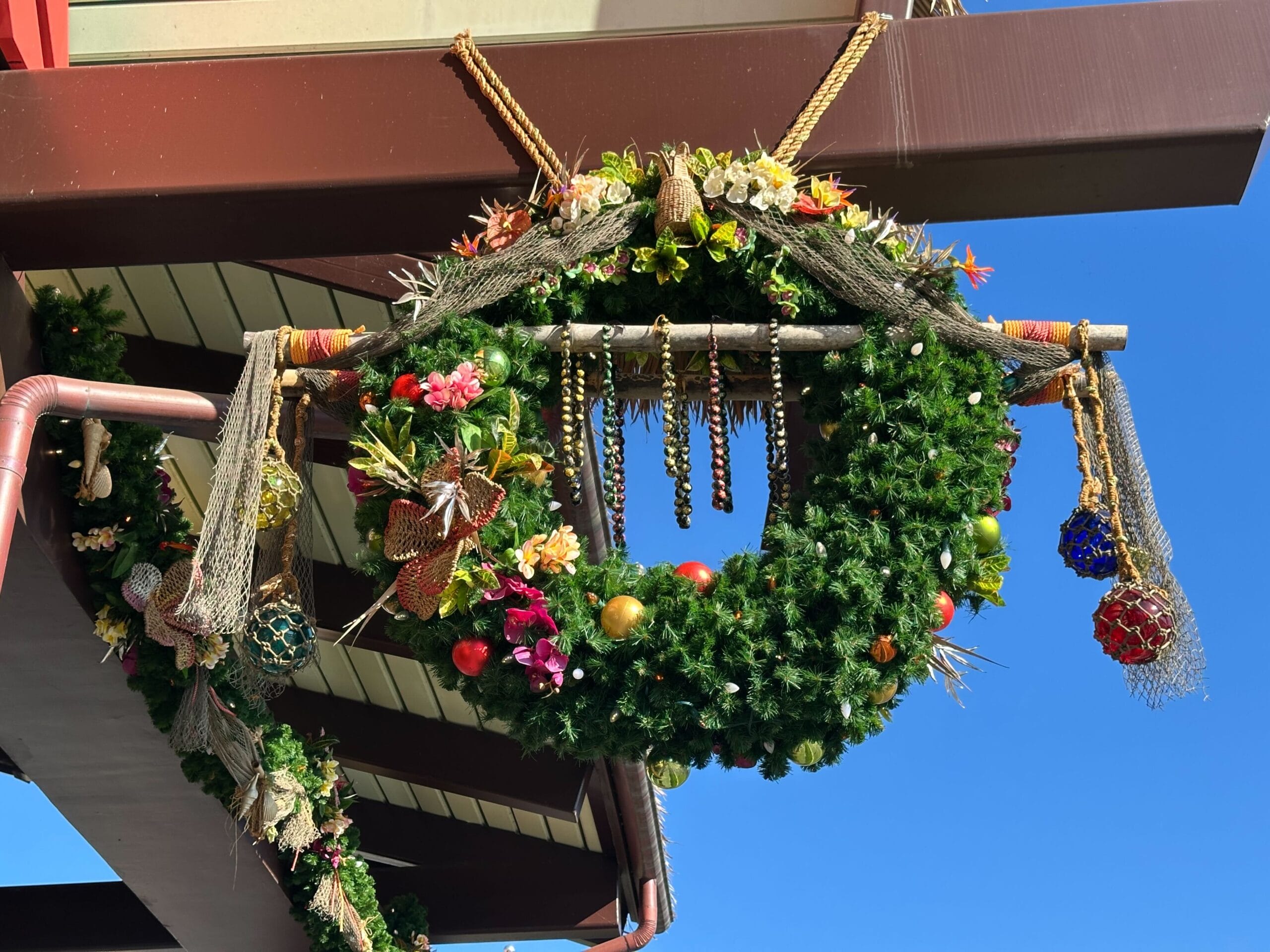 Festive holiday wreath decorated with colorful ornaments and netting, hanging from a building against a clear blue sky.