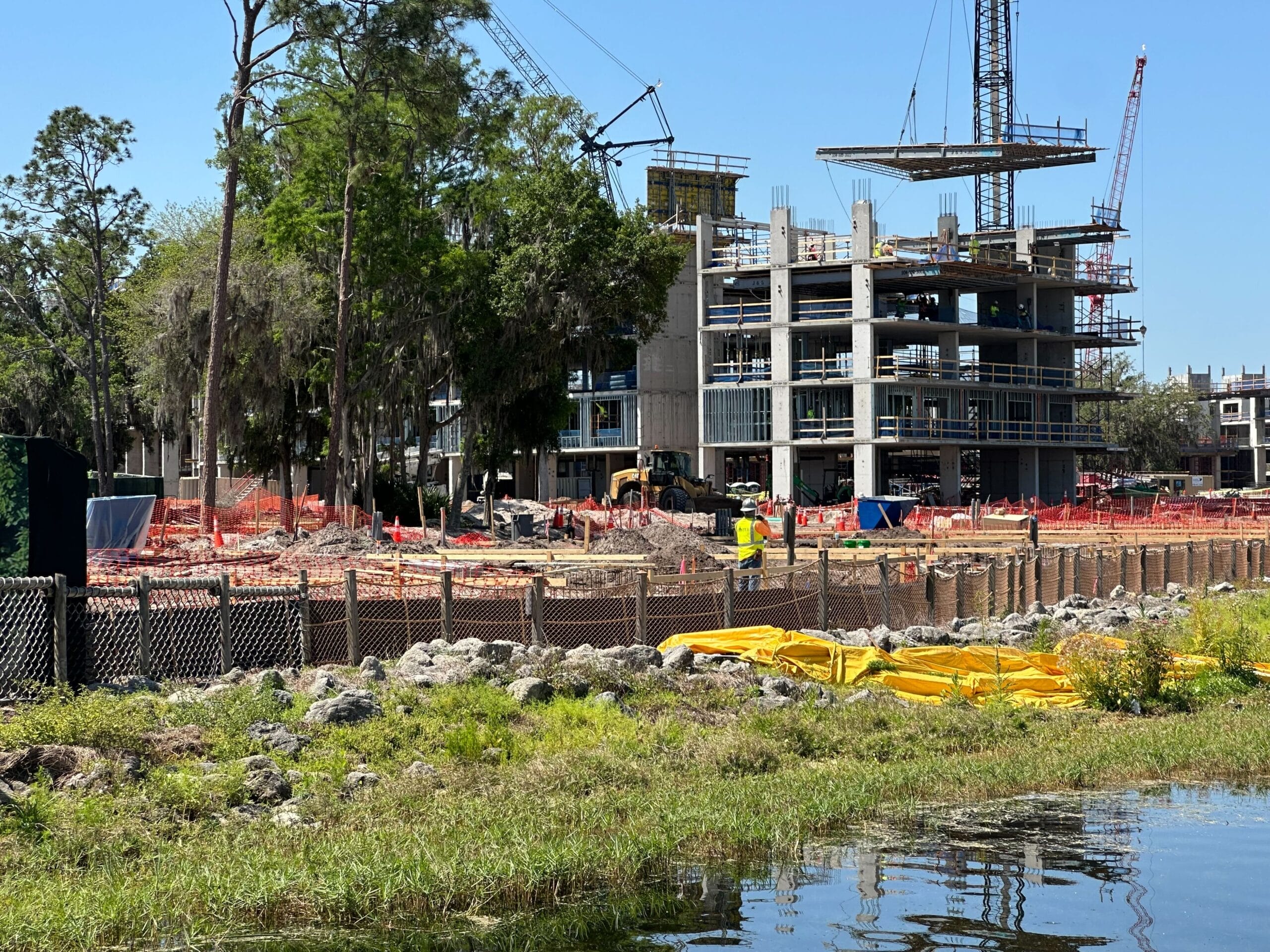 The construction site features a multi-story building framework, cranes, and materials scattered around. A fence and grassy area border the project near a serene lakeshore lodge, overlooking the water.