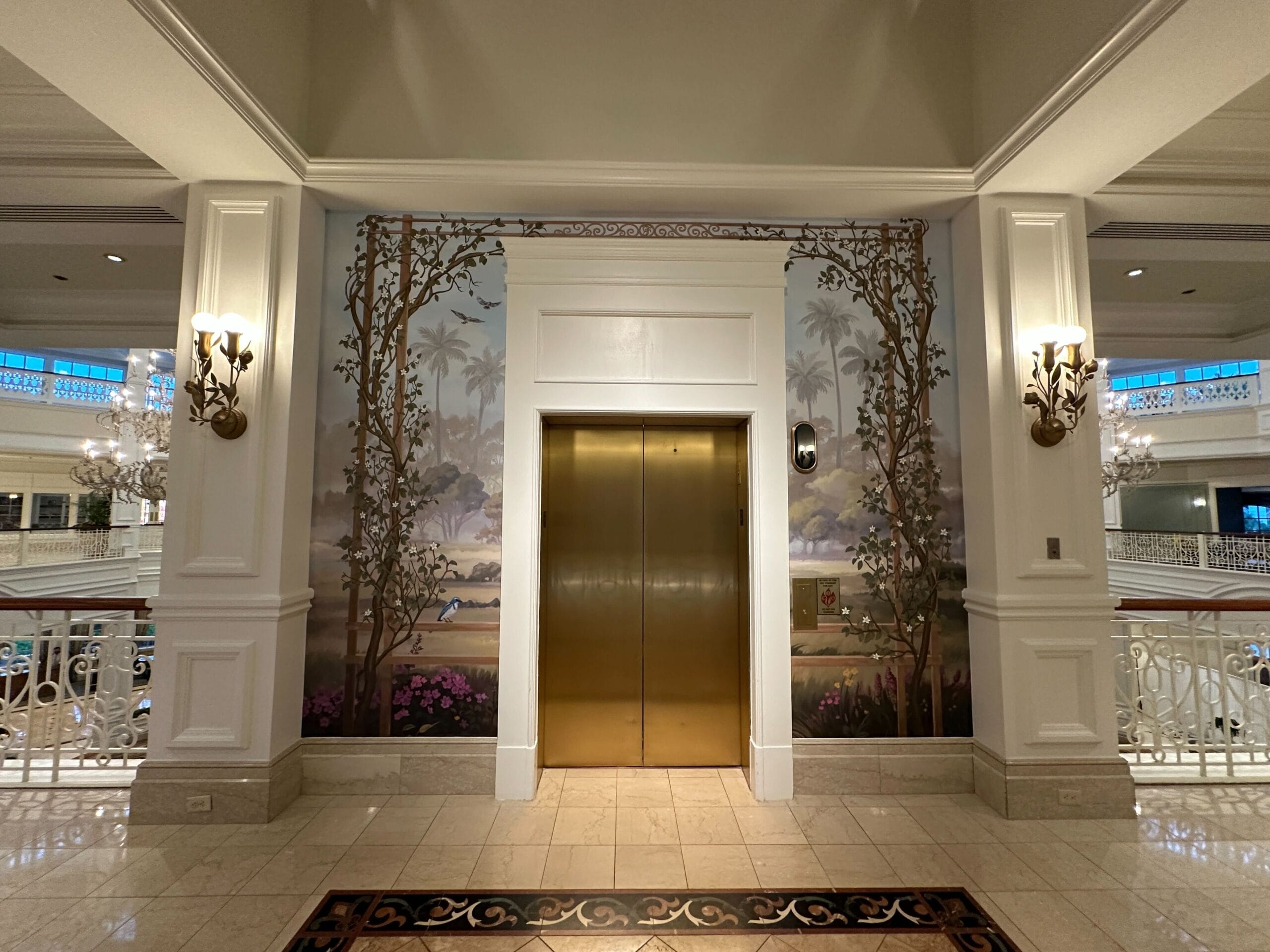 Gold elevator doors between white columns near whimsical Tea Room Windows, ornate sconces, and mural—evoking a Disney resort.
