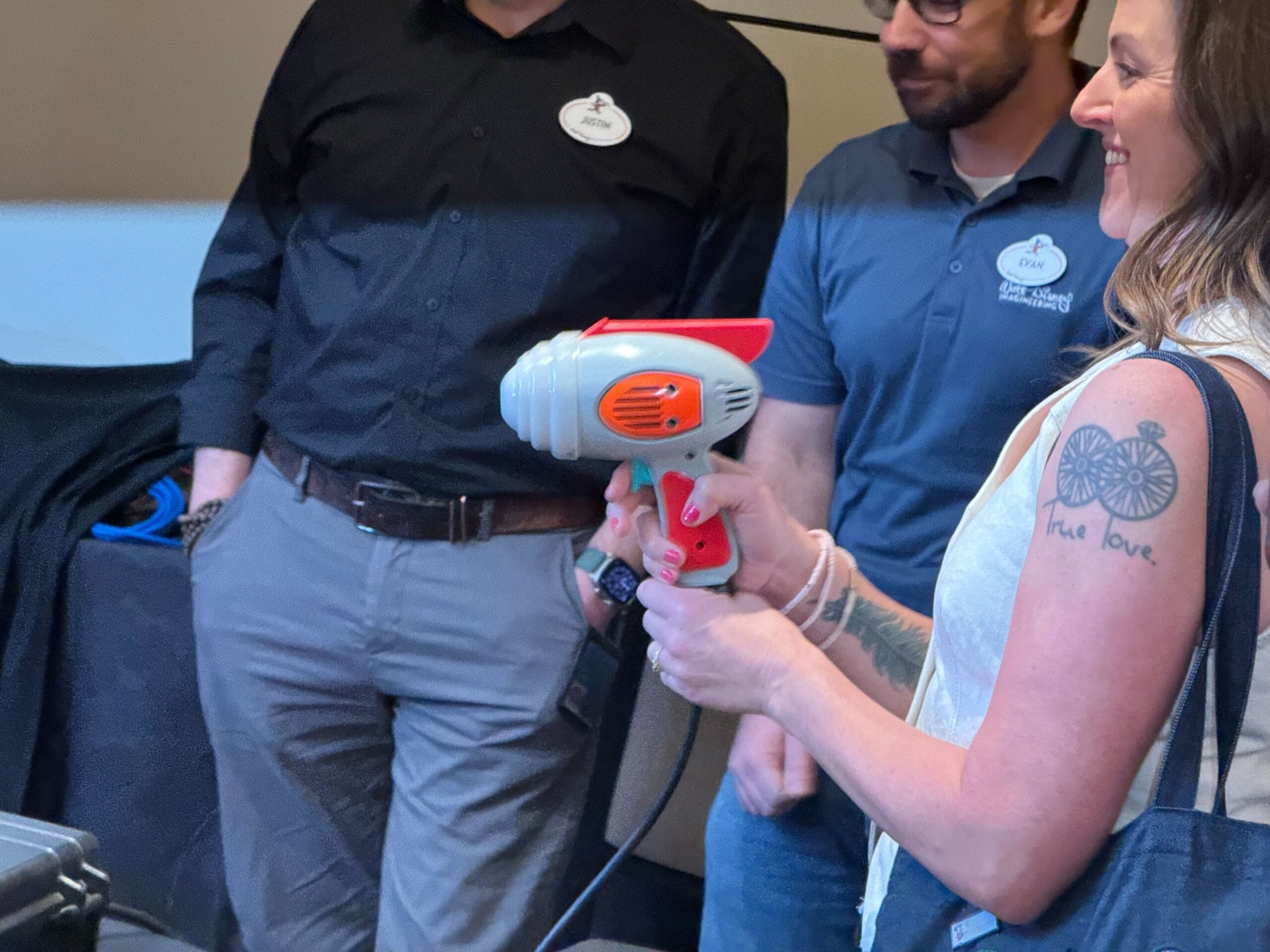 Person holding a red and gray electronic device resembling a handheld vacuum, with others observing in the background.