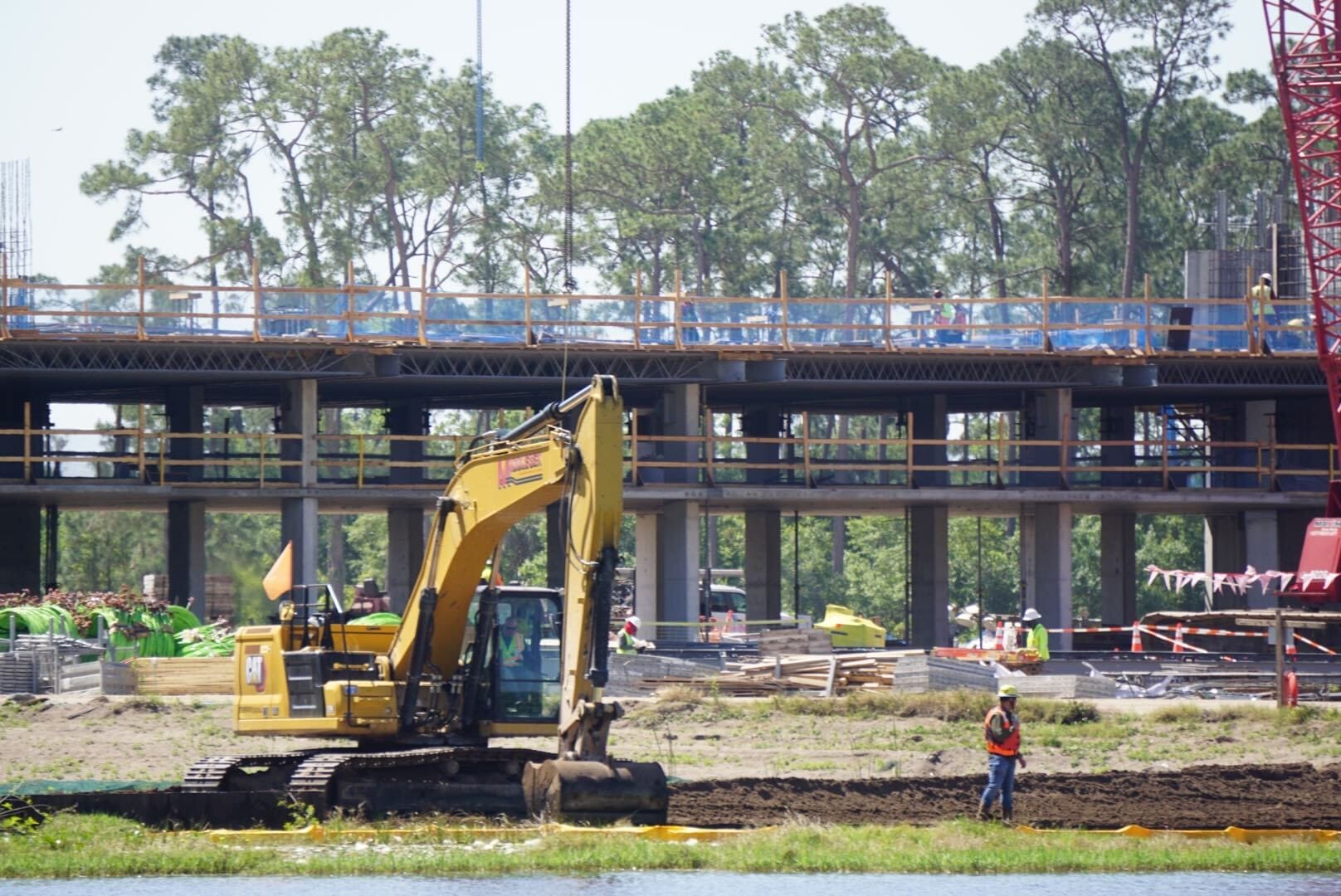 A construction site buzzes with activity as a yellow excavator and a worker in an orange vest transform the landscape. In the background, trees sway gently, hinting at the future charm of what could become the enchanting Disney Lakeshore Lodge.