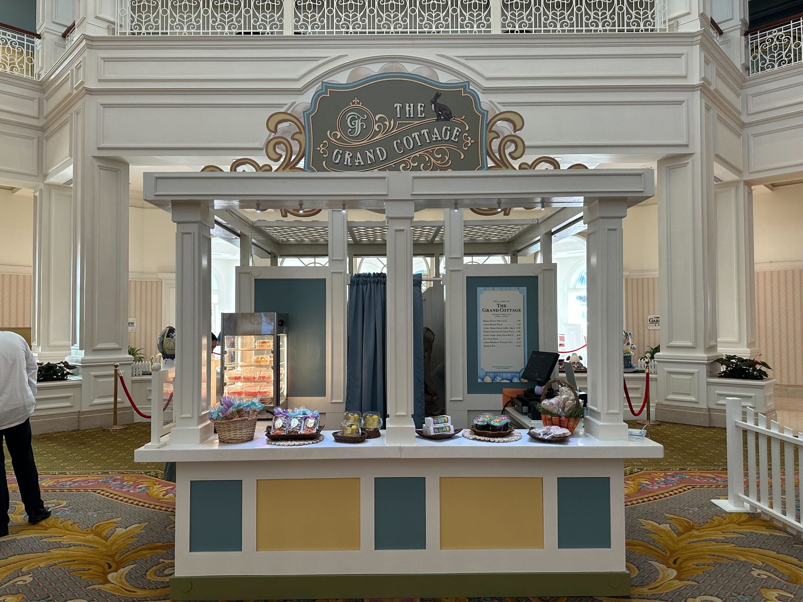 A dessert kiosk named The Grand Cottage in a lobby, featuring a display of pastries and items on the counter, with a glass cabinet and decorative sign above.